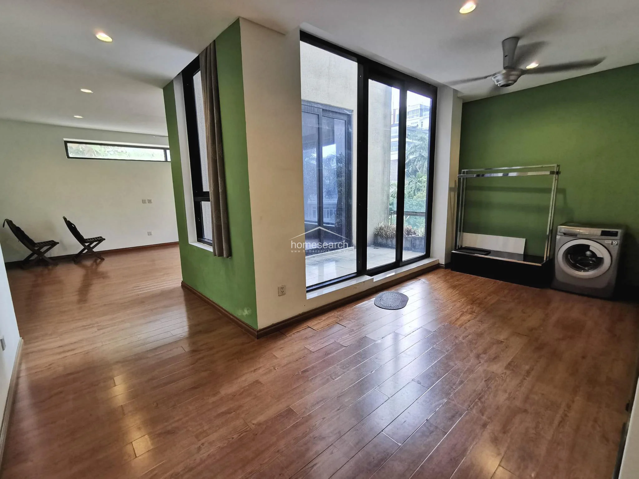 This versatile room continues the wood-look flooring theme and features a green accent wall, providing a clean and modern aesthetic. It includes a functional laundry area with a washing machine and offers direct access to a private balcony, making it a convenient and well-lit space.