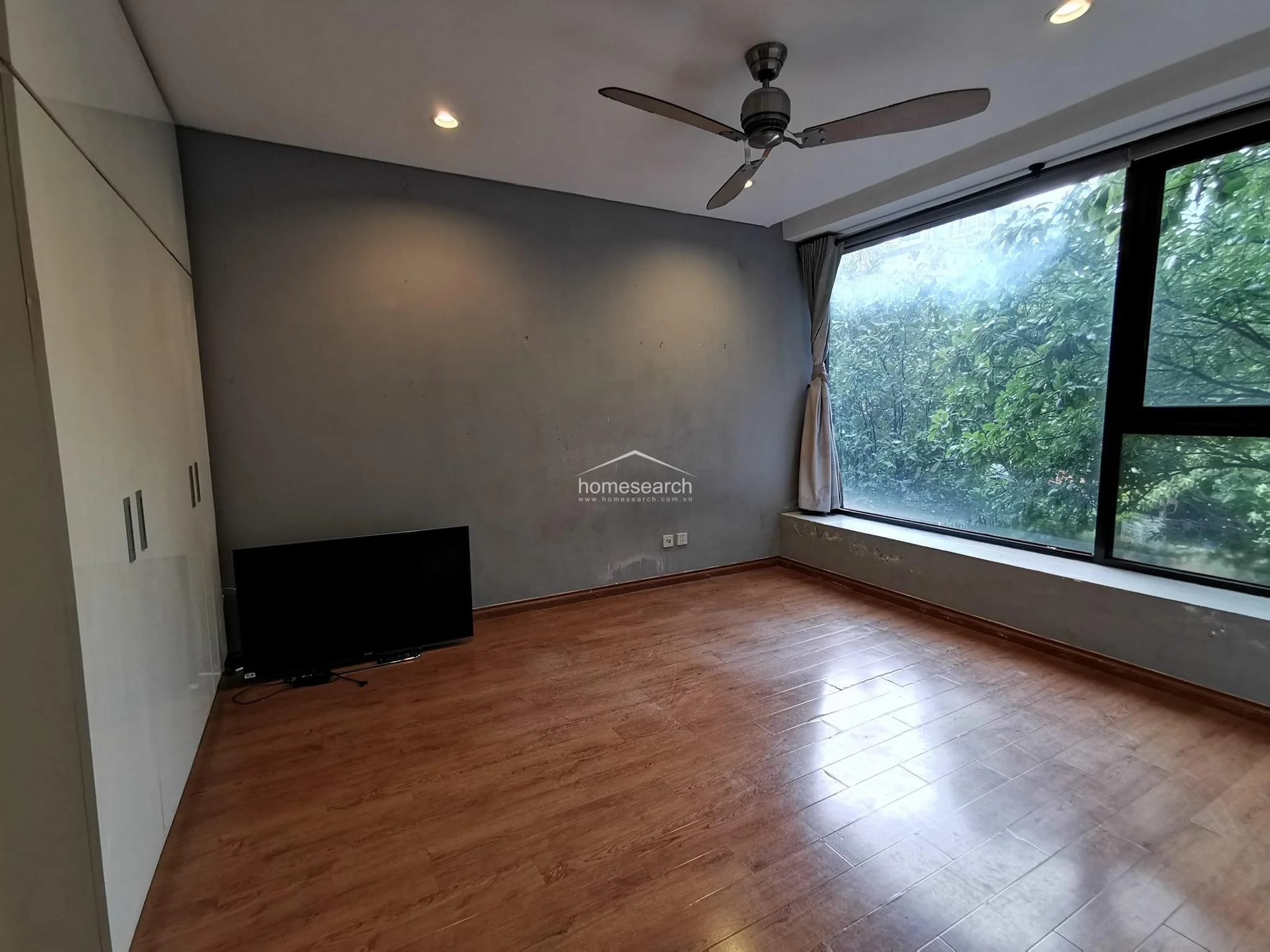 This bedroom features warm wood-look flooring and a large floor-to-ceiling window that provides an expansive view of lush greenery. The room is equipped with a ceiling fan, recessed lighting, and a sleek, white built-in wardrobe, though the grey wall shows signs of wear and requires maintenance.