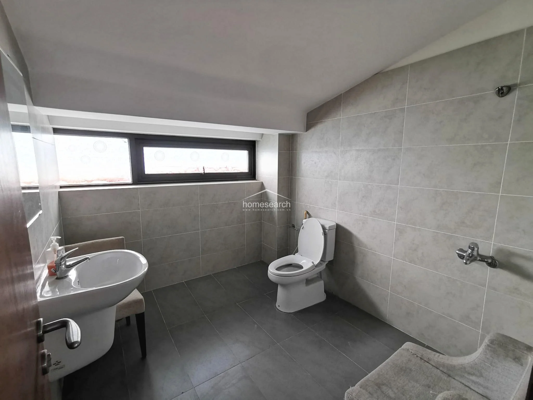 This bathroom features a minimalist design with neutral grey wall and floor tiles and a pedestal sink. It includes a large horizontal window for natural light and ventilation, though the space appears somewhat sparse and could benefit from additional fixtures or decor.