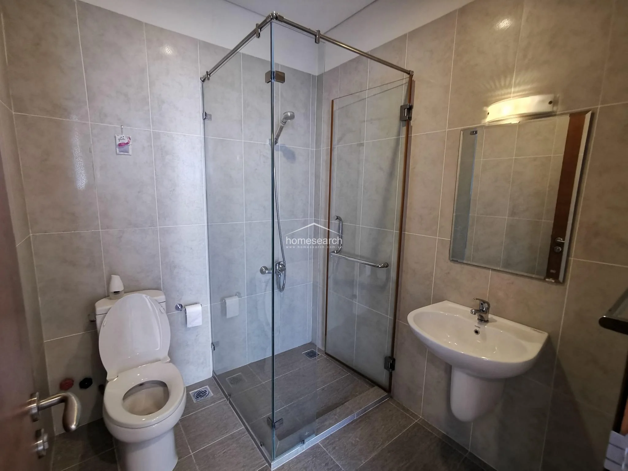 This modern bathroom is finished in neutral grey tiles and features a convenient glass-enclosed walk-in shower. The space is well-lit with recessed lighting and includes a pedestal sink and a wall-mounted mirror, presenting a clean and functional layout.