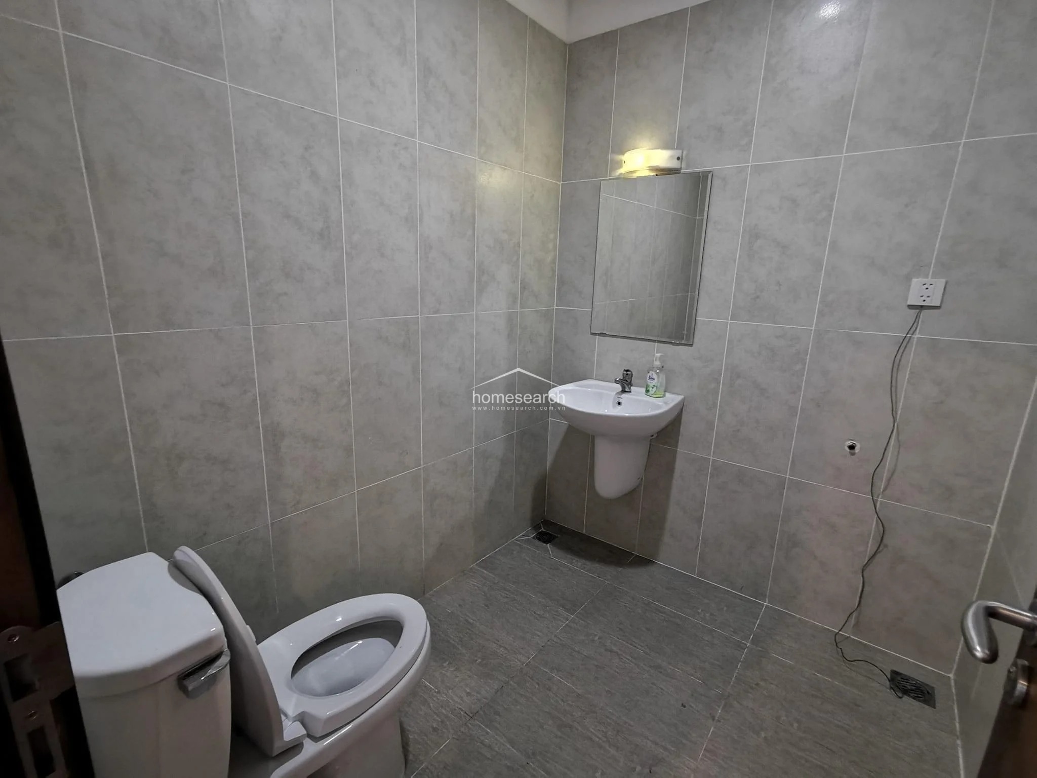 This minimalist bathroom is tiled in a neutral grey tone and features a pedestal sink with an overhead light and a simple mirror. The room is functional but basic, with a visible electrical cord suggesting a need for better cable management or additional built-in storage.