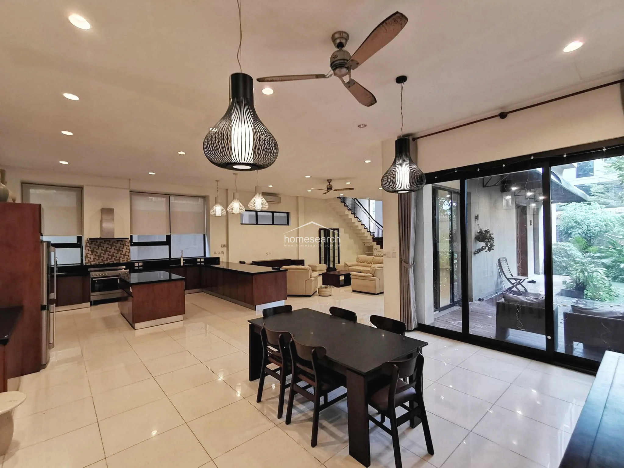 This expansive open-plan living and dining area features polished light-colored tile flooring and high ceilings with recessed lighting and decorative pendant fixtures. The layout flows effortlessly from the modern kitchen into a spacious lounge area, which connects directly to the outdoor terrace via large glass sliders. The interior is well-maintained, featuring a neutral color palette and a prominent staircase leading to the upper floor.