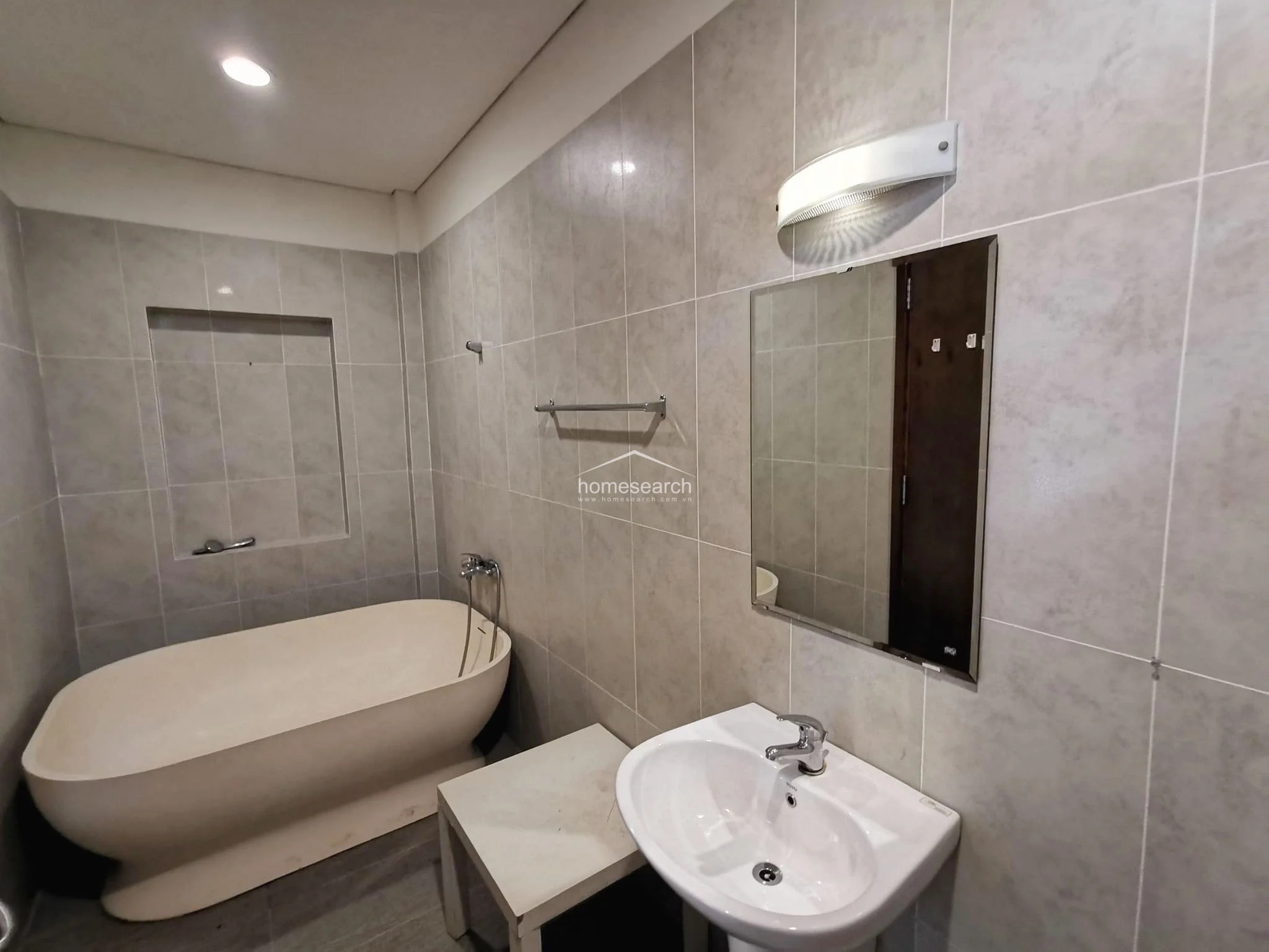 This bathroom offers a relaxing atmosphere with a freestanding soaking tub and a pedestal sink. The space is finished with neutral grey tiling, recessed lighting, and a wall-mounted mirror, providing a clean and contemporary aesthetic.