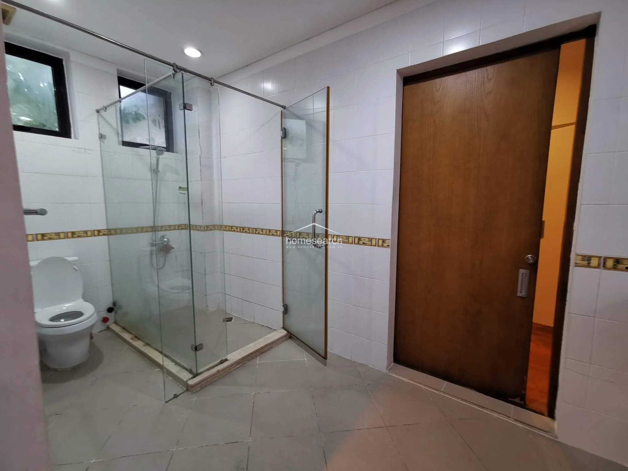 This bathroom features a walk-in glass shower enclosure with white tiled walls accented by a decorative patterned border. The space is finished with neutral floor tiles and includes a standard white toilet, with a wooden door providing access to the adjacent room.