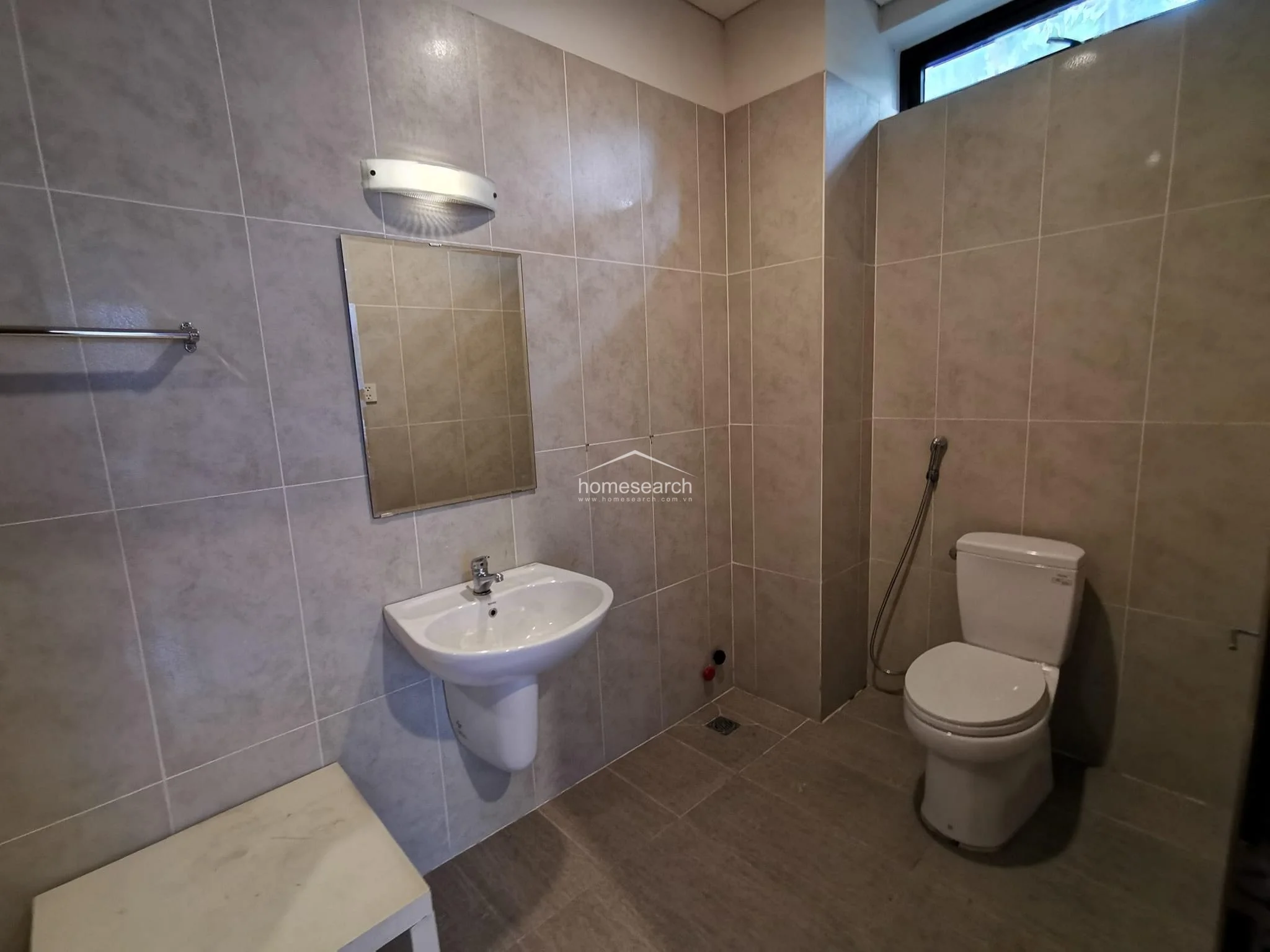 This bathroom is finished in neutral, large-format wall and floor tiles, creating a cohesive and clean aesthetic. It is equipped with a wall-mounted pedestal sink, a frameless mirror with an overhead light, and a toilet with a handheld bidet sprayer.