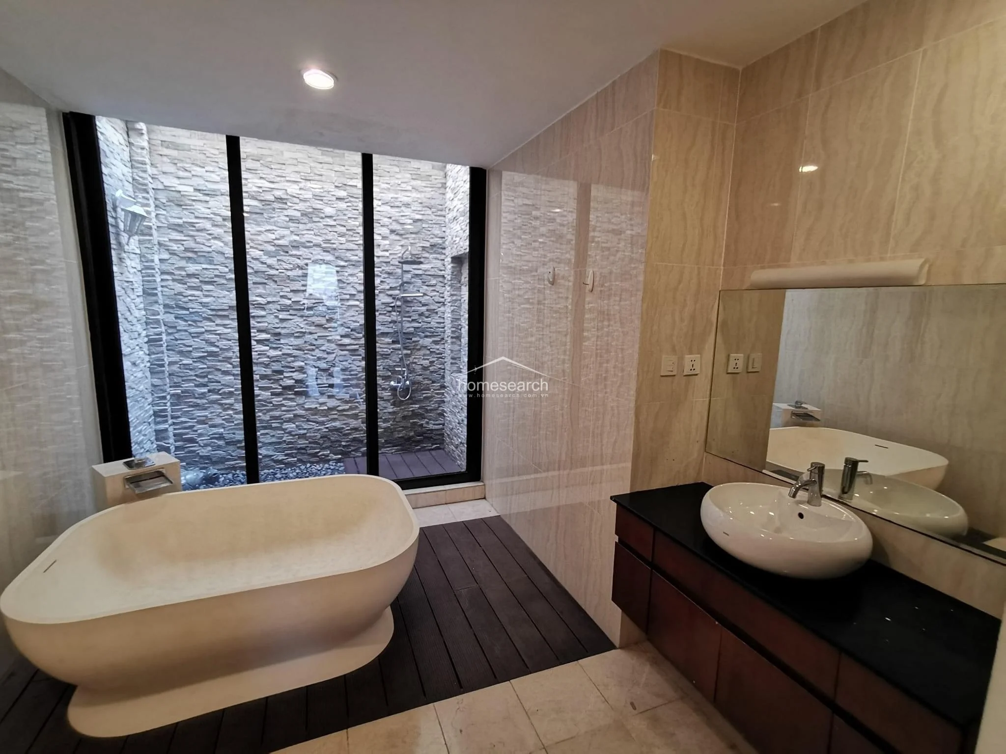 This luxurious bathroom features a freestanding soaking tub set on a dark wood-look floor, positioned next to a large floor-to-ceiling window that overlooks a private stone-walled garden area. The vanity includes a modern vessel sink on a dark countertop with wooden cabinetry, complemented by high-end wall tiling and recessed lighting.
