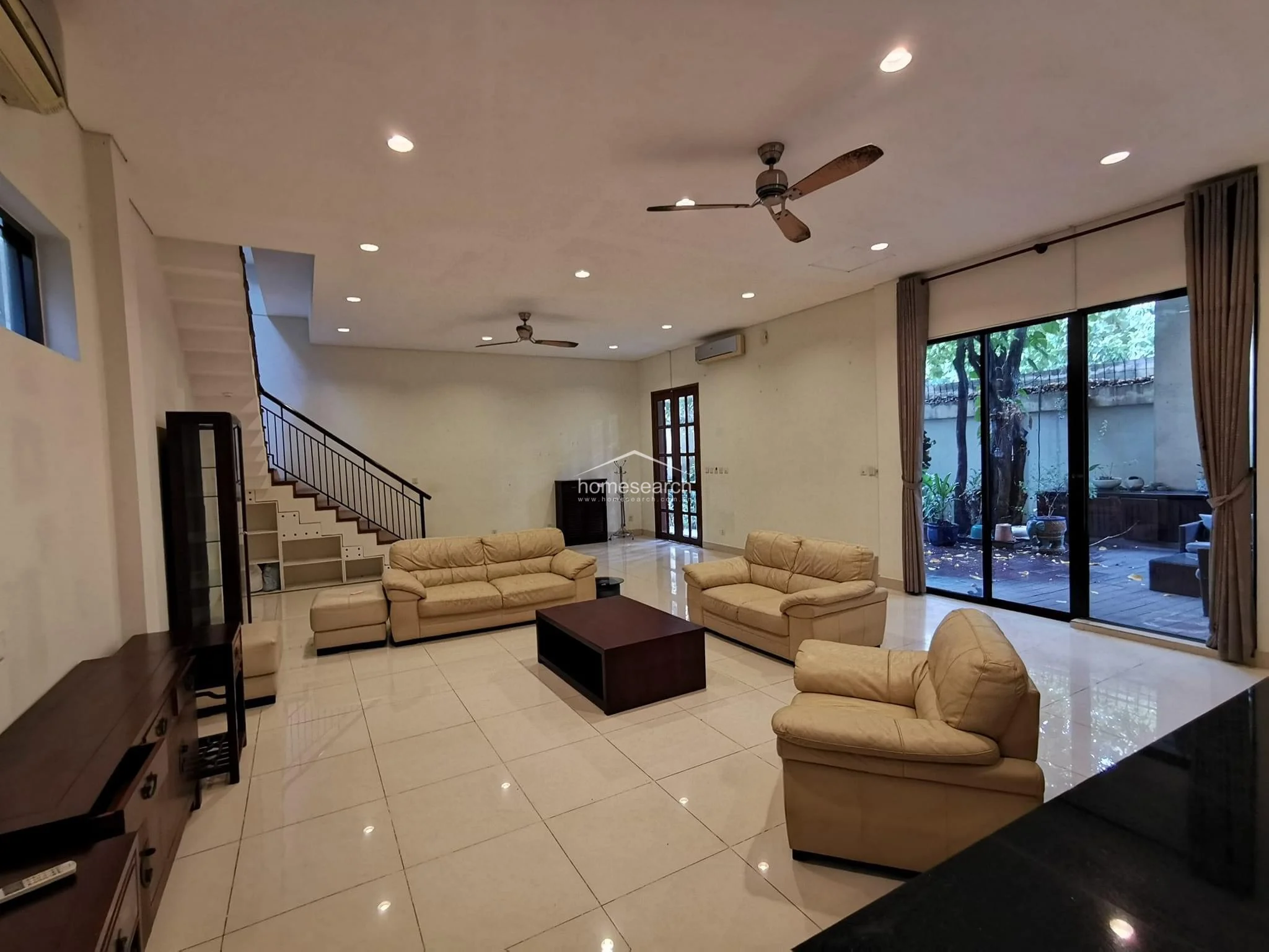 This spacious living room is anchored by a comfortable beige leather sofa set and a dark wood coffee table, creating a welcoming seating arrangement. The room benefits from excellent natural light through large glass doors that open to the garden, while the staircase adds architectural interest with its sleek metal railing. The space is clean and functional, with an air conditioning unit and ceiling fans ensuring climate control.
