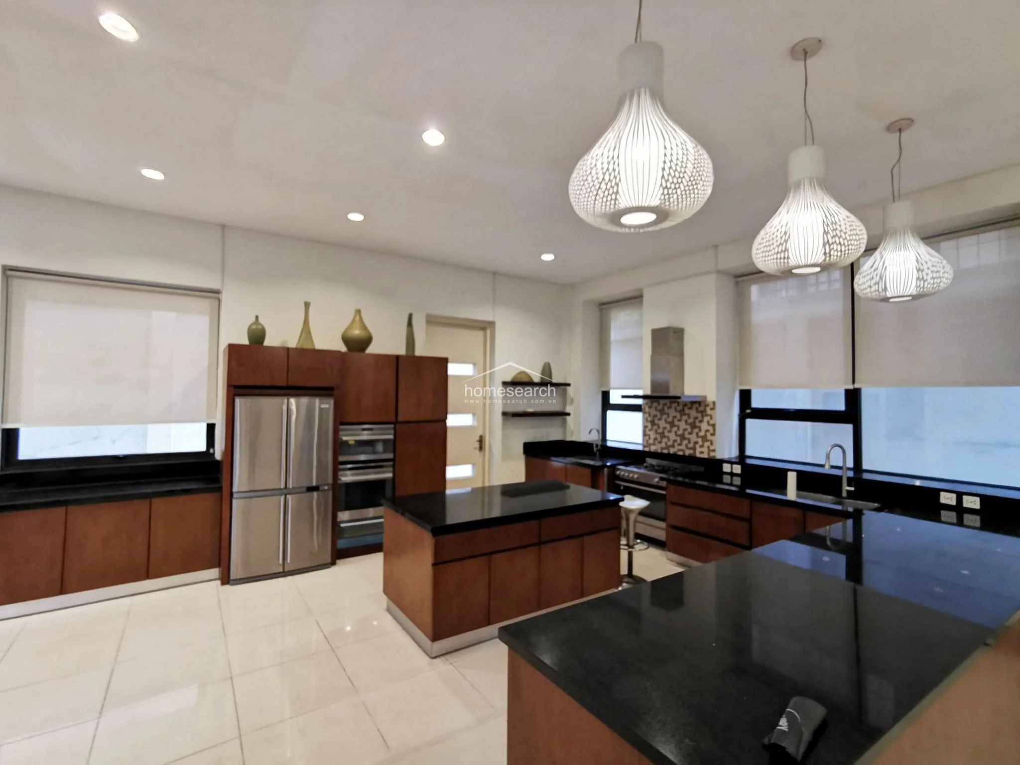 This modern kitchen is designed with a high-contrast aesthetic, featuring dark wood cabinetry paired with sleek black granite countertops and a central island. It is fully equipped with stainless steel appliances, including a double-door refrigerator and a built-in oven, and is illuminated by stylish, contemporary pendant lights. The layout is highly functional with ample counter space and storage, complemented by large windows that provide bright natural light.