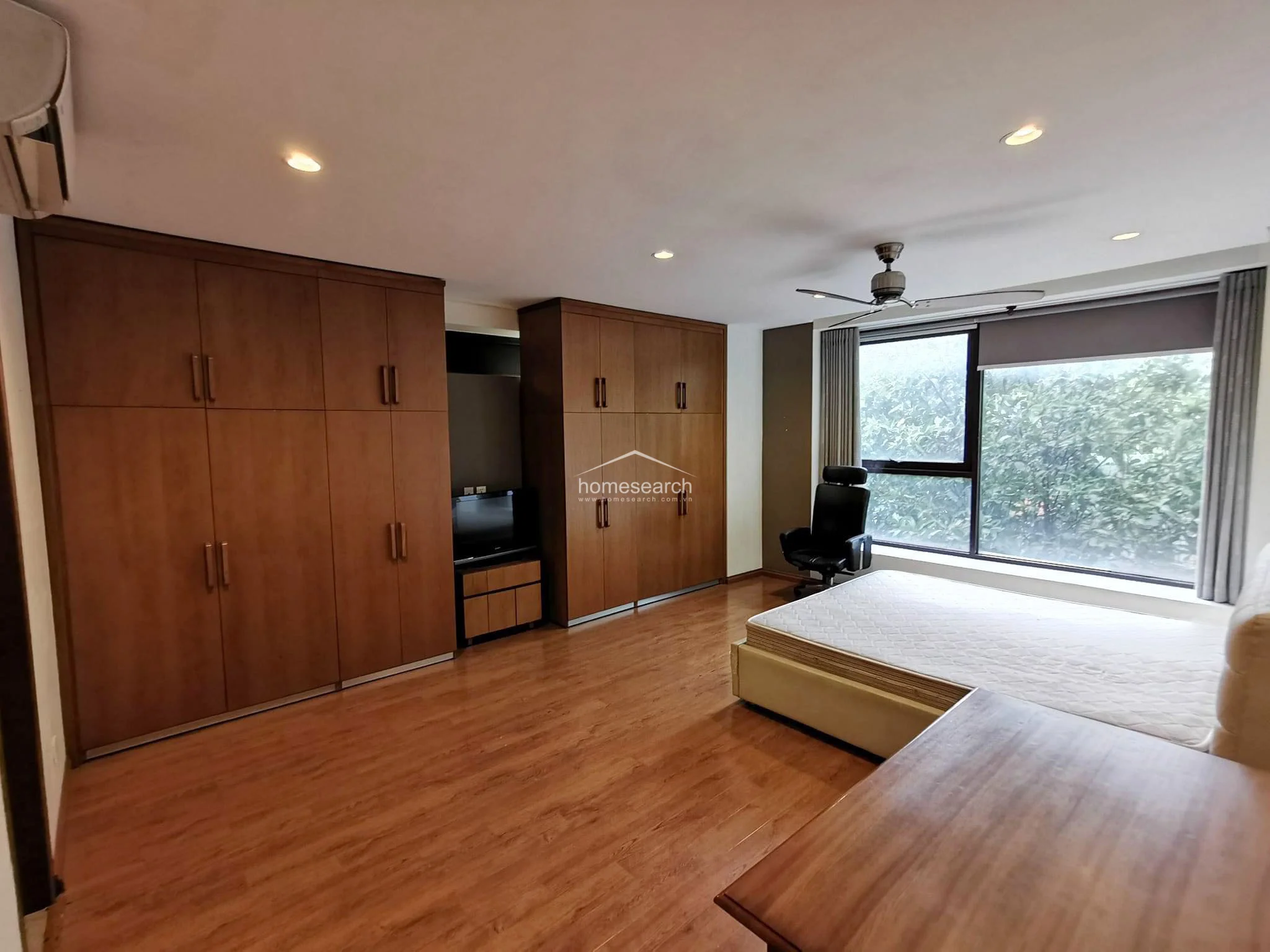 This bedroom features warm wood-look flooring and extensive floor-to-ceiling wooden built-in wardrobes that offer generous storage capacity. A large window provides ample natural light and a pleasant green view, complemented by a ceiling fan, recessed lighting, and a minimalist aesthetic.
