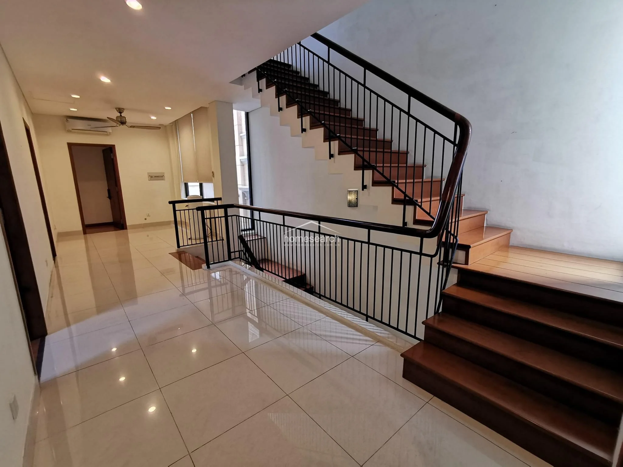This transitional hallway area showcases a striking architectural staircase with dark wood treads and a sleek black metal railing that connects the different levels of the home. The space maintains a clean, modern look with light-colored tile flooring, recessed ceiling lights, and minimalist wall finishes.