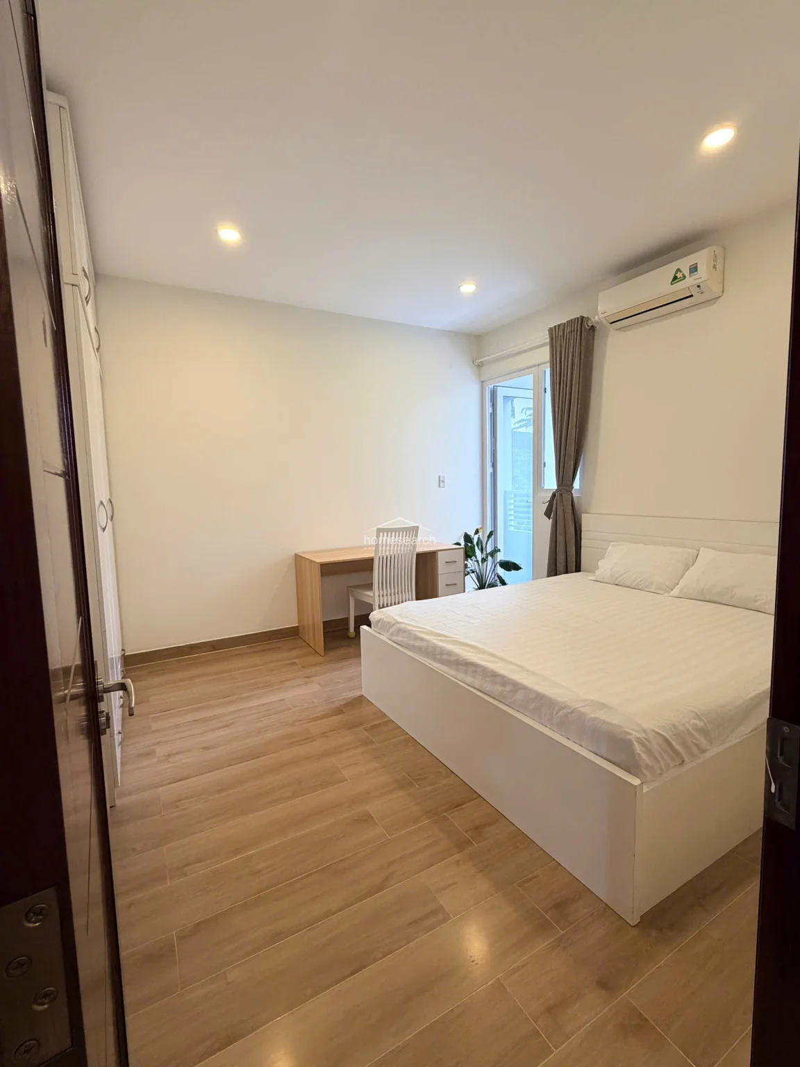 This bedroom offers a bright and airy atmosphere, highlighted by a glass door that provides access to an outdoor balcony. A dedicated wooden desk and chair create a convenient workspace, complemented by a white bed frame and neutral grey curtains.
