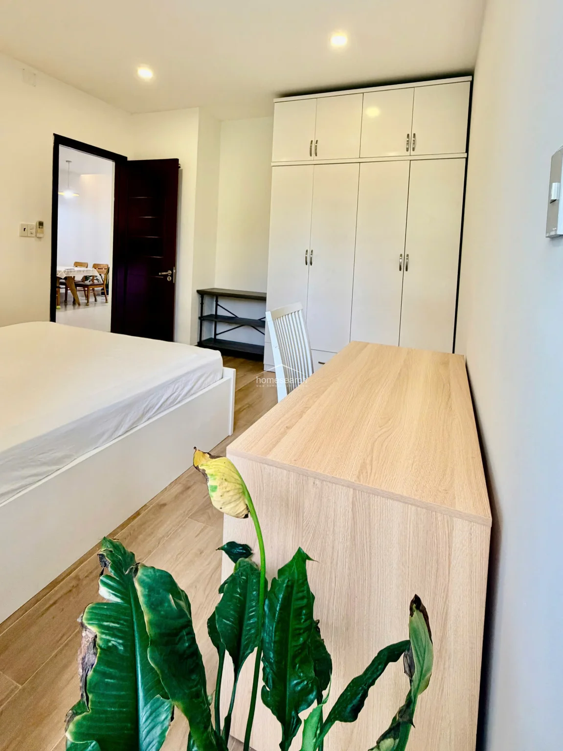 This bedroom features a clean, minimalist aesthetic with light wood-look flooring and neutral white walls. It includes a floor-to-ceiling white wardrobe for ample storage, a matching desk, and a direct line of sight into the adjacent dining area through a dark-framed doorway.