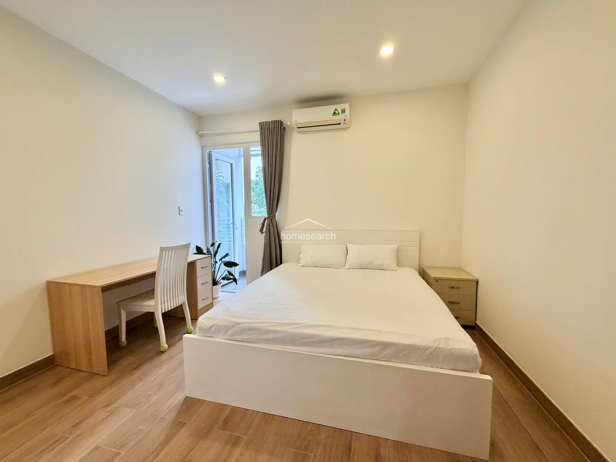 This view emphasizes the bedroom's efficient use of space, featuring a light wood desk and a matching bedside table. The room maintains a cohesive, modern look with white furnishings, recessed lighting, and a neutral color palette throughout.