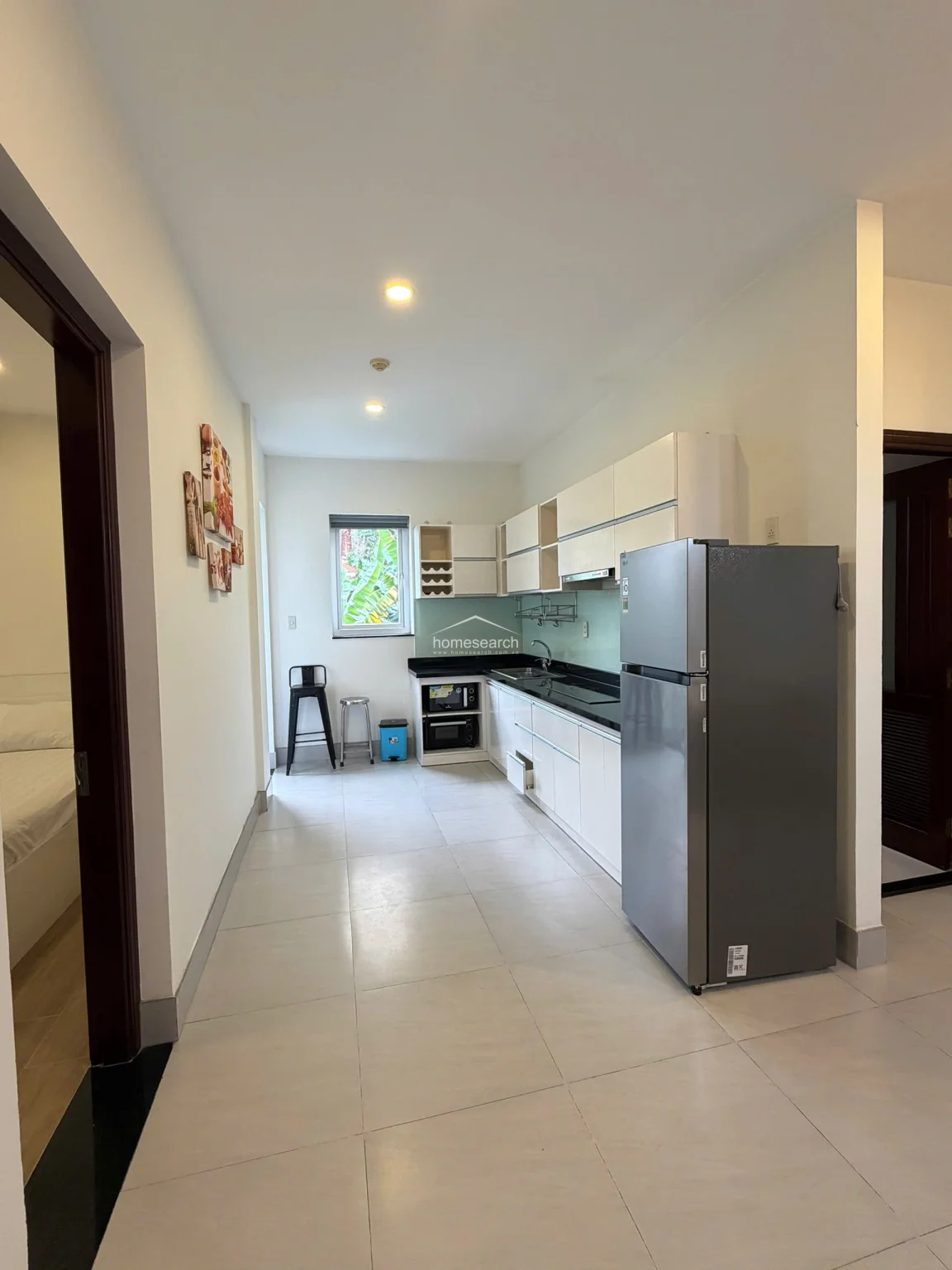 The kitchen is designed in a linear, efficient layout with white cabinetry, black countertops, and a light green glass backsplash. It is well-equipped with a stainless steel refrigerator, a built-in cooktop, and an under-cabinet microwave, while the large window ensures natural light flows into the workspace.