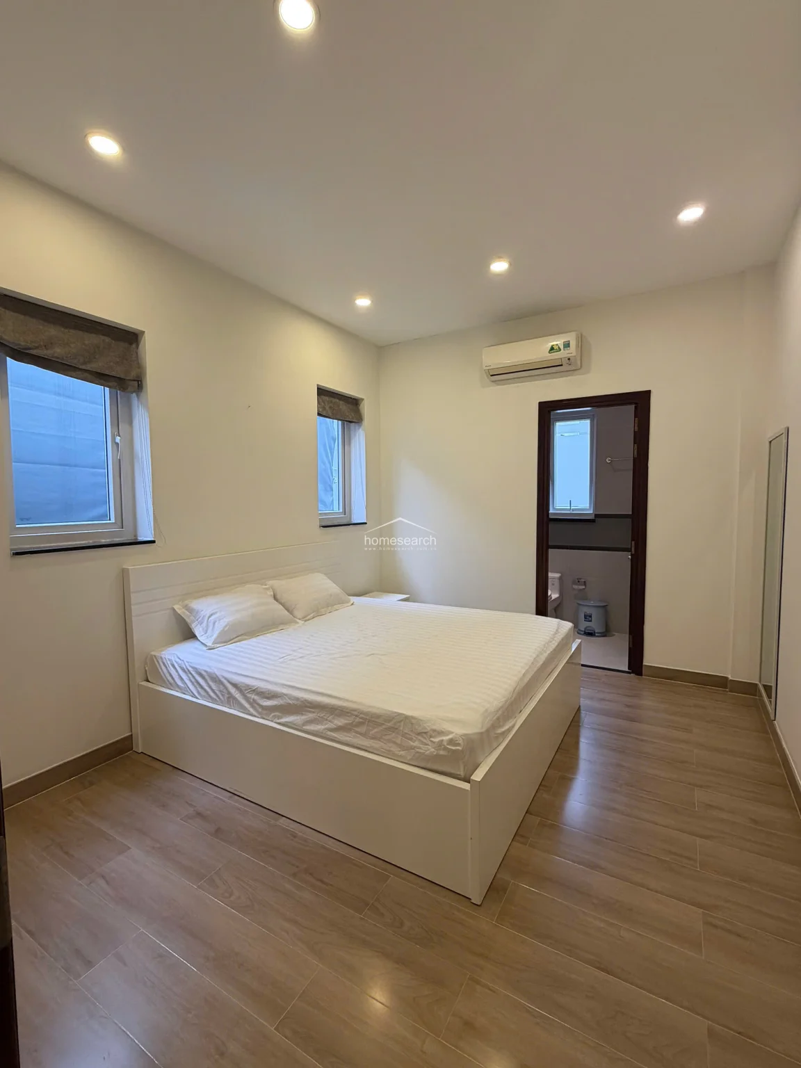 This bedroom is well-lit by two windows and includes a wall-mounted AC unit for climate control. The room connects directly to an en-suite bathroom, featuring a simple, modern aesthetic with consistent wood-look flooring and recessed ceiling lights.