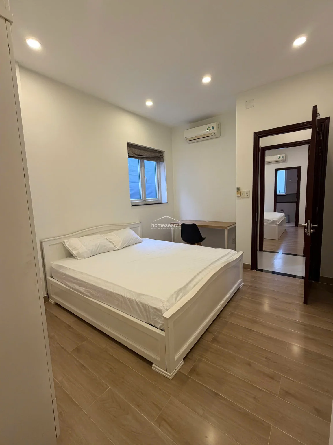 This bedroom features warm wood-look flooring and a simple, modern design centered around a white bed frame and a small workspace. The room is connected to the rest of the unit via a doorway that reveals a glimpse of another bedroom, and it includes a wall-mounted AC unit for climate control.