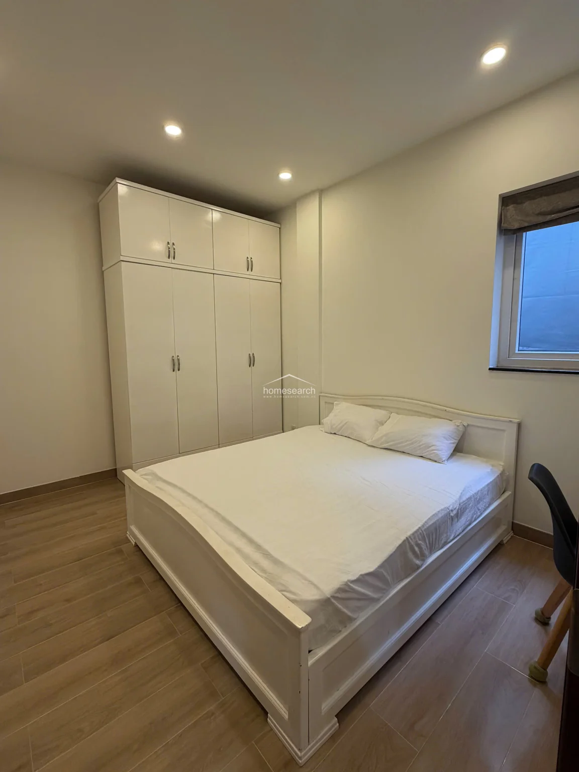 This bedroom is anchored by a large, white floor-to-ceiling wardrobe that maximizes vertical storage. The space is kept simple and functional, with a white bed frame, wood-look tile flooring, and a single window that provides natural light.