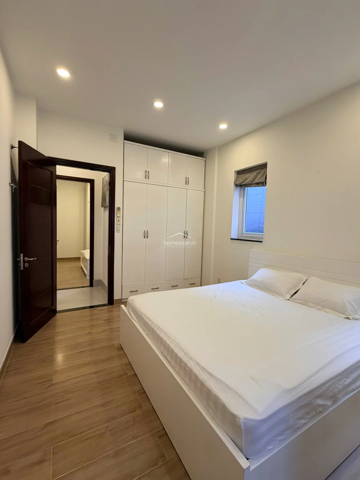 This bedroom features a clean, minimalist design with light wood-look tile flooring and crisp white walls. A large, floor-to-ceiling white wardrobe provides ample storage, while a dark wood door leads to an adjacent room, suggesting a functional layout.