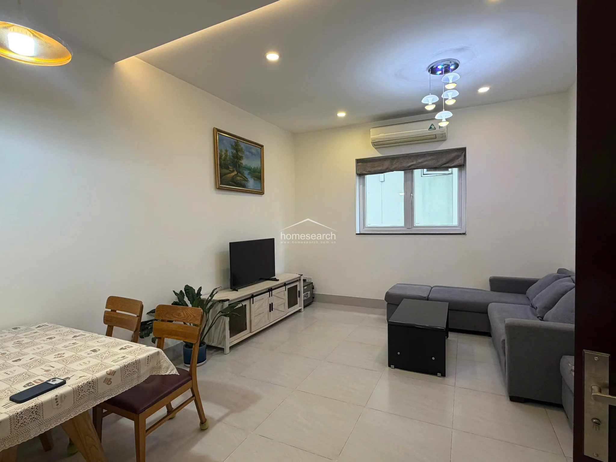 This living area features a clean, minimalist layout with light-colored tile flooring and white walls that create a bright, airy atmosphere. The space is furnished with a grey sectional sofa, a wooden TV console, and a dining set, all illuminated by recessed ceiling lights and a decorative chandelier.