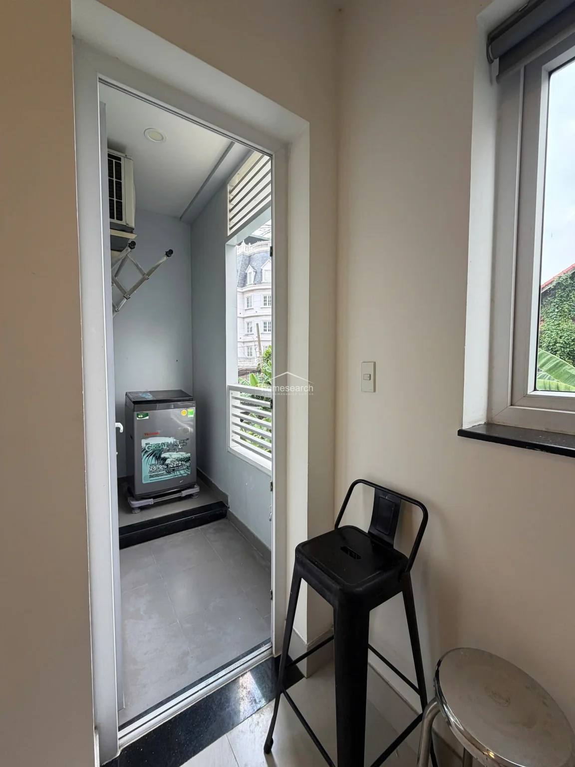 This utility space functions as a laundry balcony, featuring a Toshiba washing machine and a wall-mounted drying rack. The area is finished with grey floor tiles and offers a view of neighboring architecture, while a modern black metal stool sits just inside the interior room threshold.