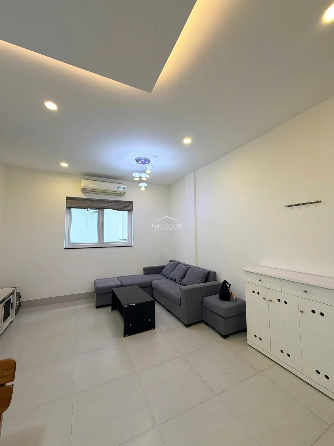 This view highlights the functional living room layout, showing the connection to the adjacent dining area and the inclusion of a wall-mounted AC unit. The room maintains a consistent aesthetic with neutral tones, large-format floor tiles, and a white storage cabinet that provides practical organization.