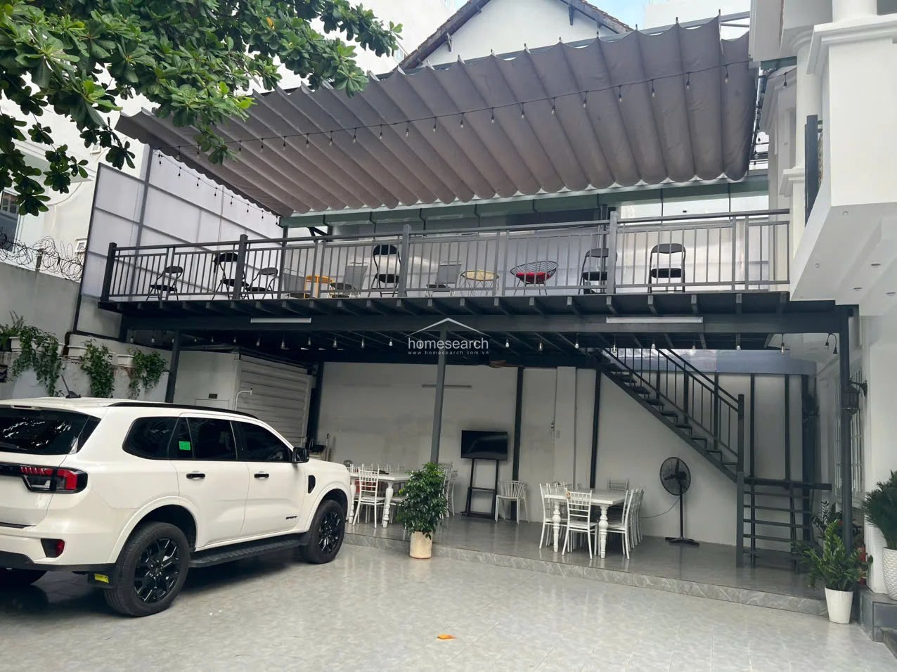 This outdoor space features a spacious paved driveway and parking area, complemented by a covered patio with dining tables and a ceiling fan for comfortable al fresco gatherings. A large, retractable fabric awning provides shade for the upper-level balcony, which is equipped with modern metal railings and outdoor seating.