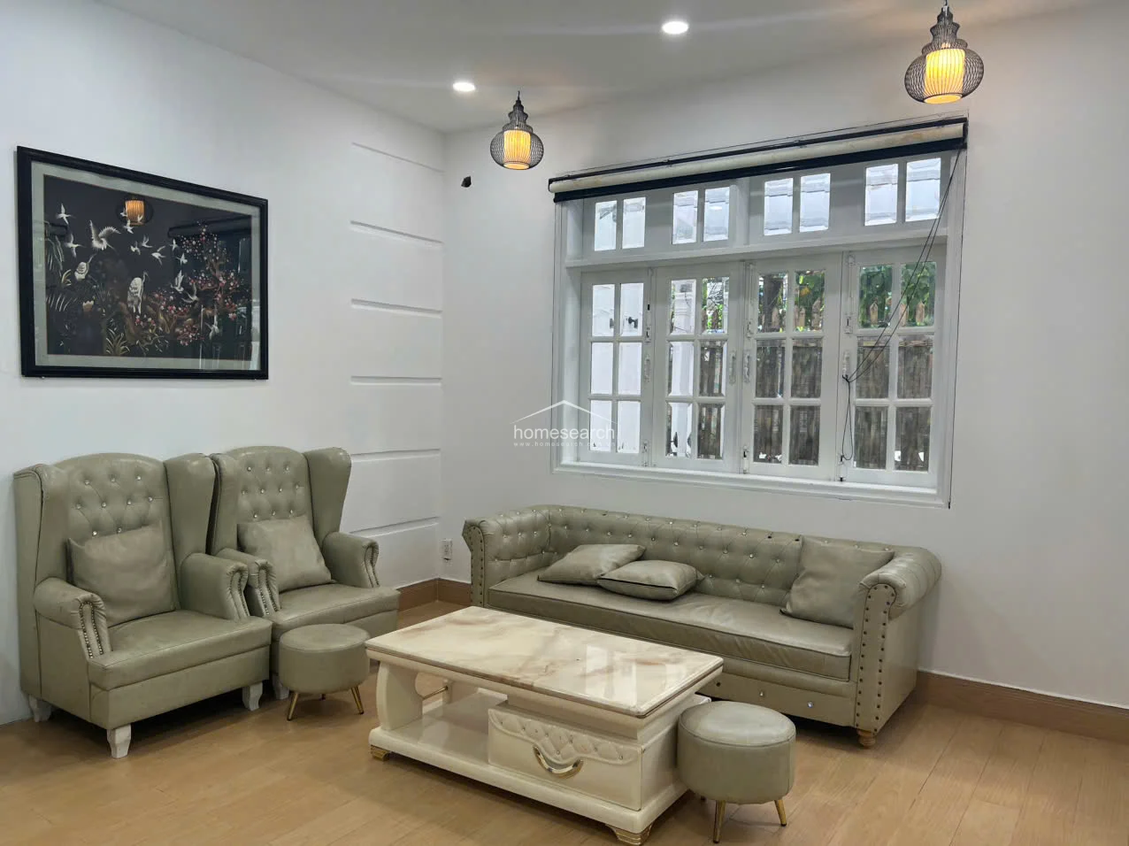 This living area is finished with light wood-look flooring and crisp white walls, centered around a classic tufted leather sofa set and a matching marble-top coffee table. Natural light streams in through a large, multi-pane window, while elegant pendant lights and a framed piece of wall art add a touch of sophistication to the room.