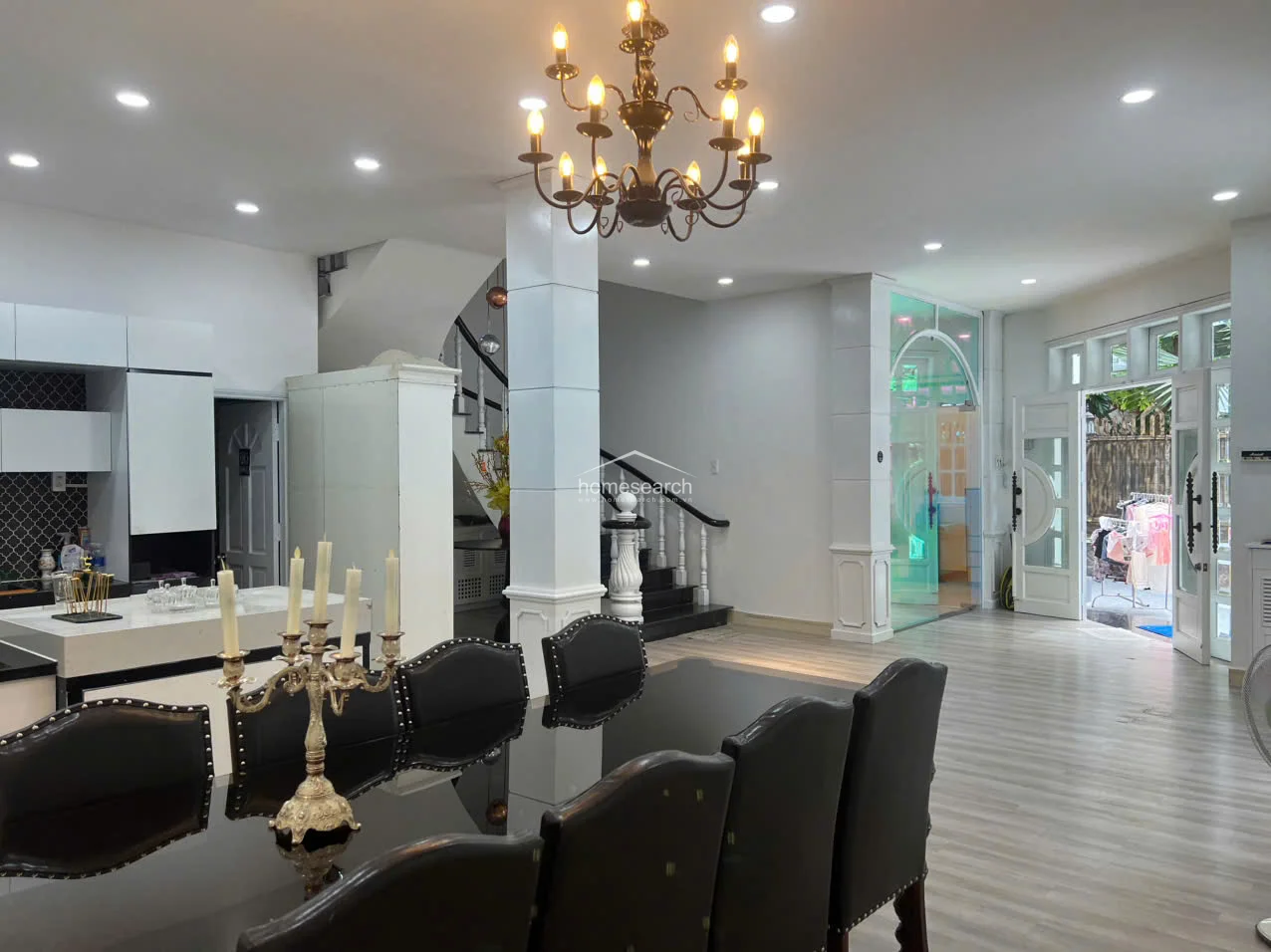 The open-plan dining and kitchen area showcases a striking black dining set beneath a traditional chandelier, creating a formal atmosphere. The space flows seamlessly toward the exterior through glass-paneled double doors, while the kitchen is anchored by a modern island with a stone countertop and integrated stovetop.