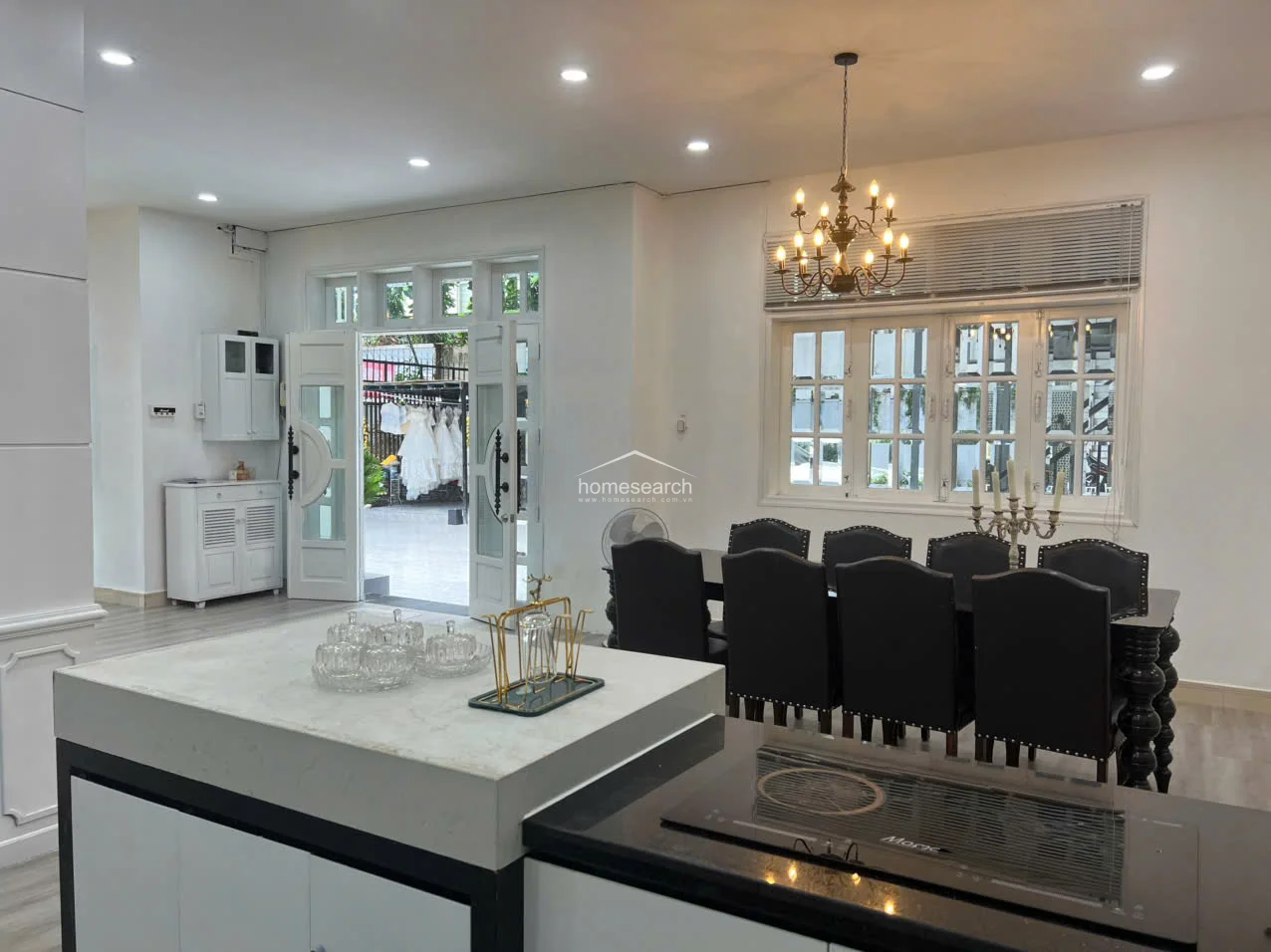 This view highlights the kitchen's functional layout, featuring a sleek island with a marble-style countertop and a built-in induction cooktop. The dining area remains connected to the kitchen, and the room is well-lit by recessed ceiling lights and a large, multi-pane window that overlooks the outdoor area.
