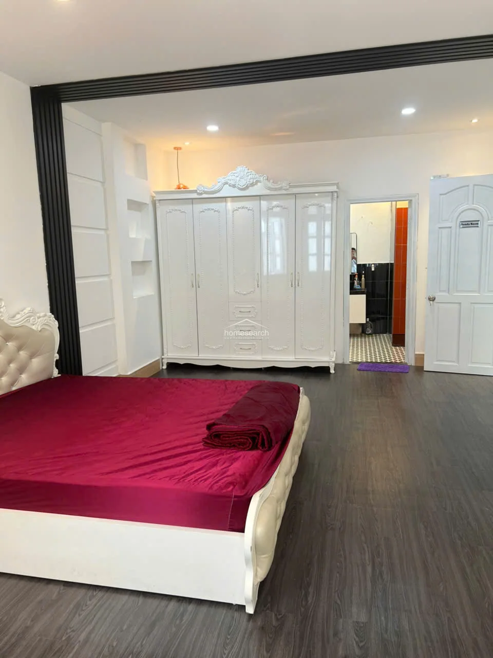This bedroom features dark wood-toned flooring and a prominent white ornate wardrobe that provides ample storage space. The room includes a tufted bed frame with deep red bedding and offers direct access to a private en-suite bathroom with a distinctive tiled floor.