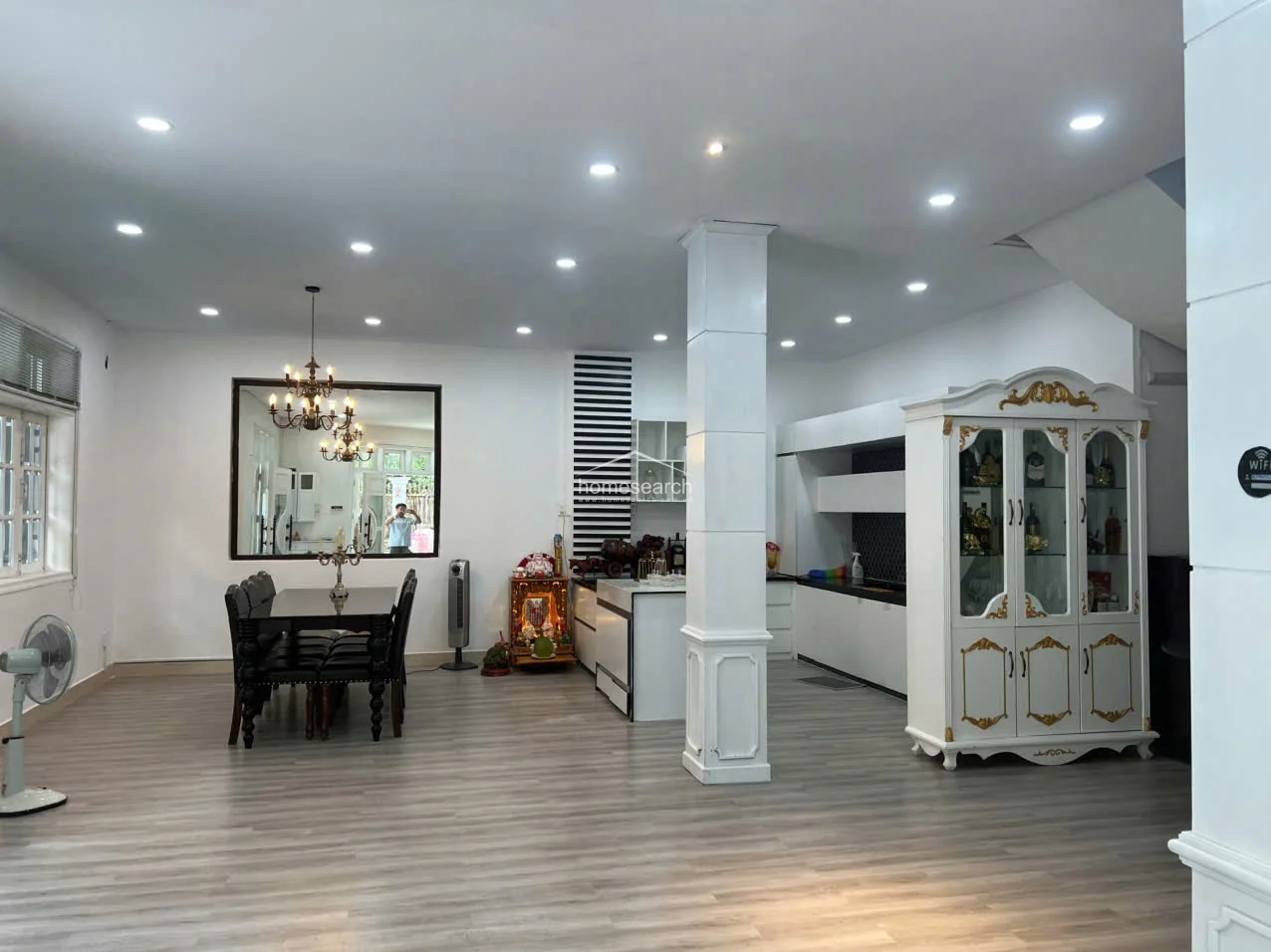 This open-concept living and dining area features light wood-look flooring and a clean, white color scheme accented by a striking black-and-white striped wall feature. The space is anchored by a prominent structural column and includes a formal dining set, a decorative display cabinet, and recessed ceiling lighting that creates a bright, modern atmosphere.