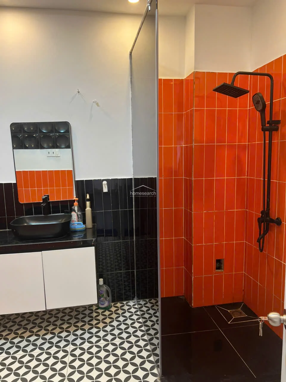 This bathroom features a bold, high-contrast design with black-and-white patterned floor tiles, black wall tiles, and a vibrant orange tiled shower surround. It is equipped with a modern black rainfall showerhead, a sleek black vessel sink on a white vanity, and a glass partition to separate the wet and dry zones.