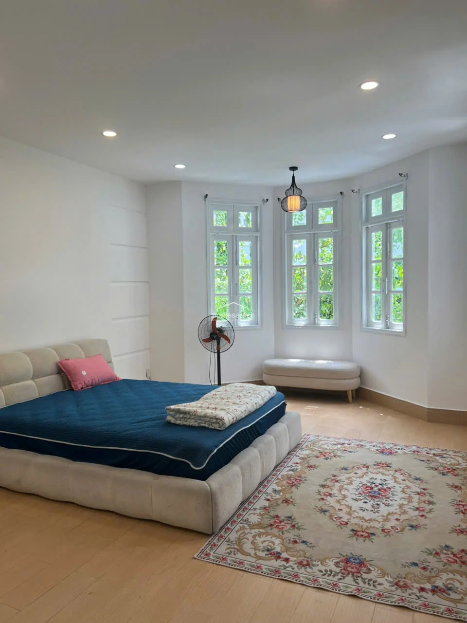 This spacious bedroom is bathed in natural light from a large bay window, which offers a pleasant view of greenery and includes a built-in window bench. The room features light wood flooring, neutral white walls, and a contemporary upholstered bed frame, all contributing to a clean and minimalist aesthetic.