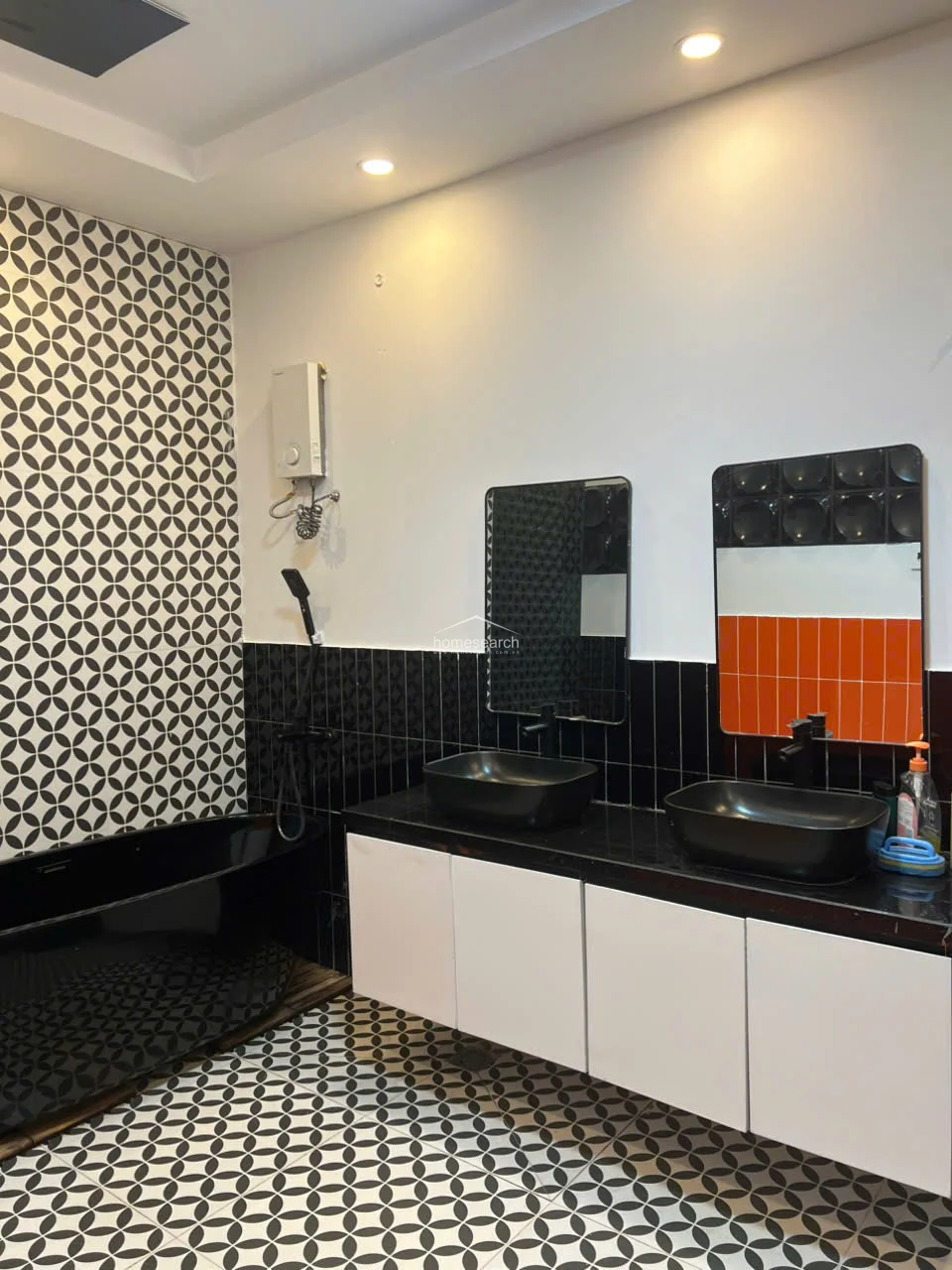 This modern bathroom showcases a sophisticated design with a black freestanding bathtub and a double-sink vanity featuring black vessel sinks and a black countertop. The room is finished with a striking combination of geometric patterned floor tiles, black wall tiles, and a splash of orange, complemented by recessed lighting and a wall-mounted water heater.