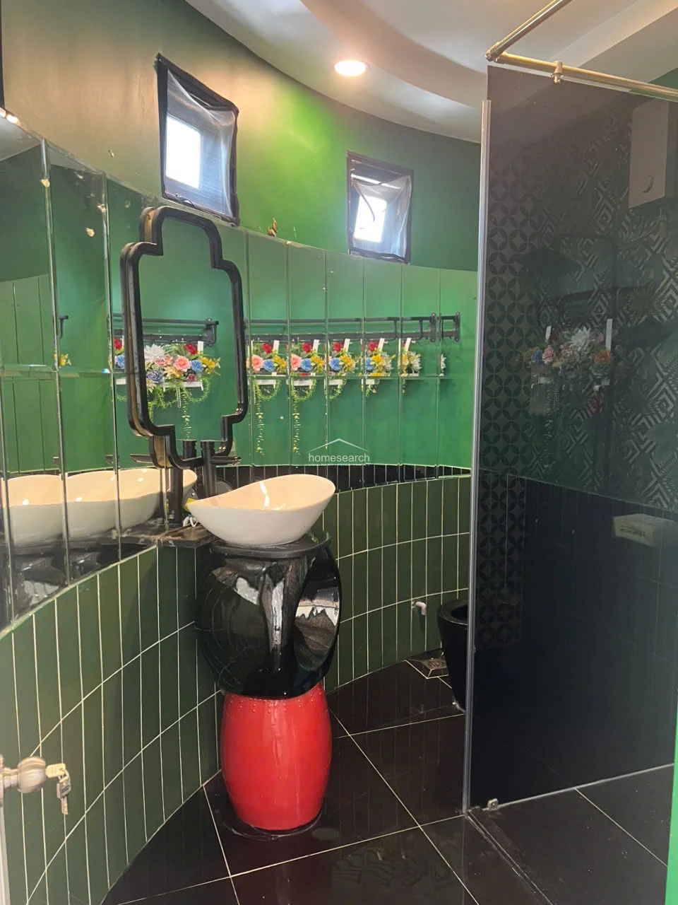 This uniquely designed bathroom features a vibrant green color scheme with emerald-tiled walls, a curved mirror arrangement, and a distinctive pedestal sink with a red base. The space includes a glass-enclosed shower area with patterned privacy glass and high-end fixtures, reflecting a bold and artistic approach to interior design.