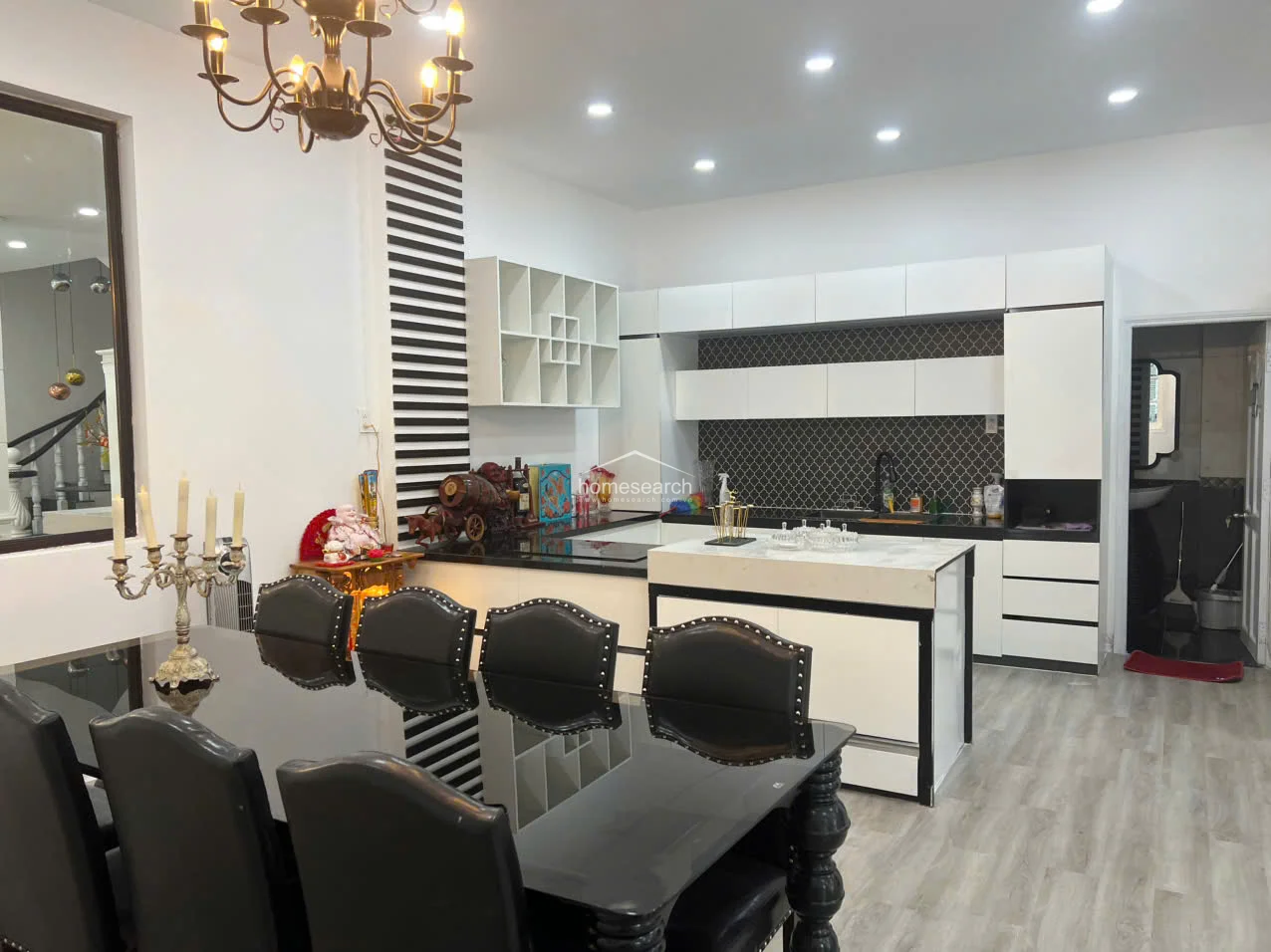 This open-concept dining and kitchen area features a modern, high-contrast black and white color scheme with light wood-look flooring. The space is anchored by a formal black dining set and a kitchen island with a marble-style countertop, illuminated by recessed lighting and a traditional chandelier.