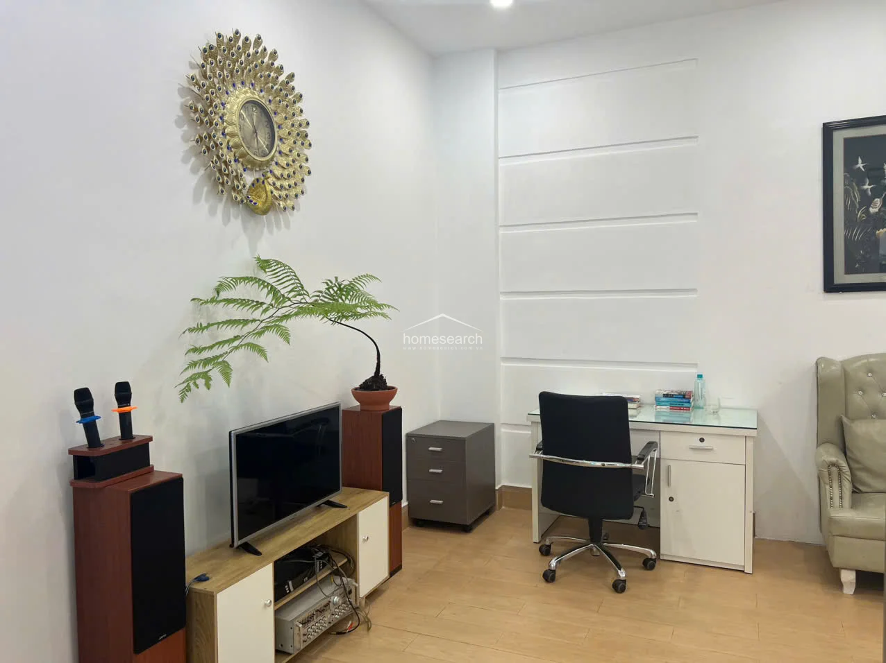 This home office or study area features light wood-tone flooring and crisp white walls accented by a decorative, peacock-inspired wall clock. The space is furnished with a modern white desk, a black ergonomic office chair, and a compact media console, maintaining a clean and organized atmosphere.