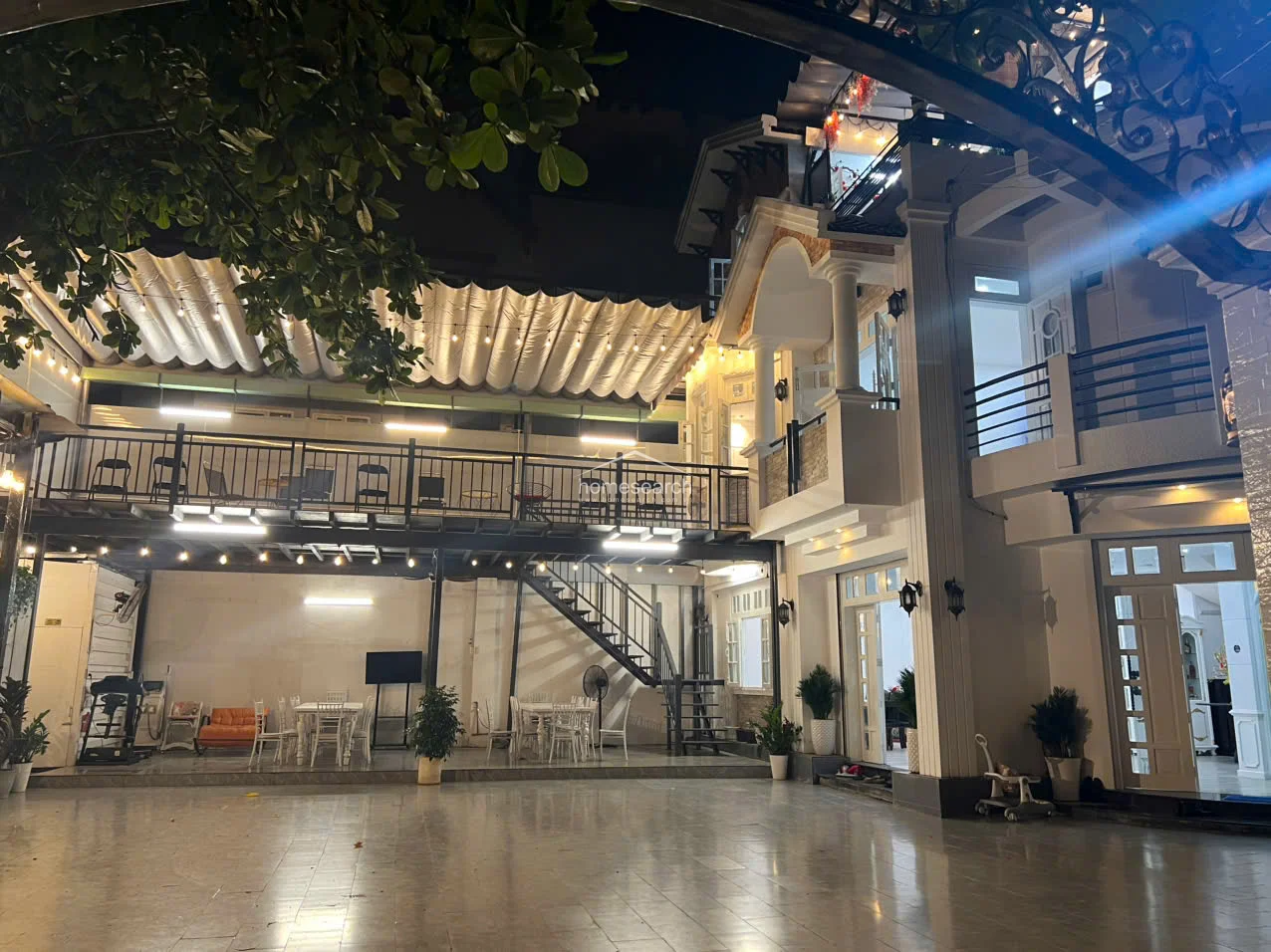 This expansive outdoor courtyard serves as a multi-functional living and entertainment space, featuring a large tiled patio and a covered upper-level balcony with string lighting. The architectural design incorporates classical columns, arched entryways, and a modern retractable awning system, all maintained in excellent condition.