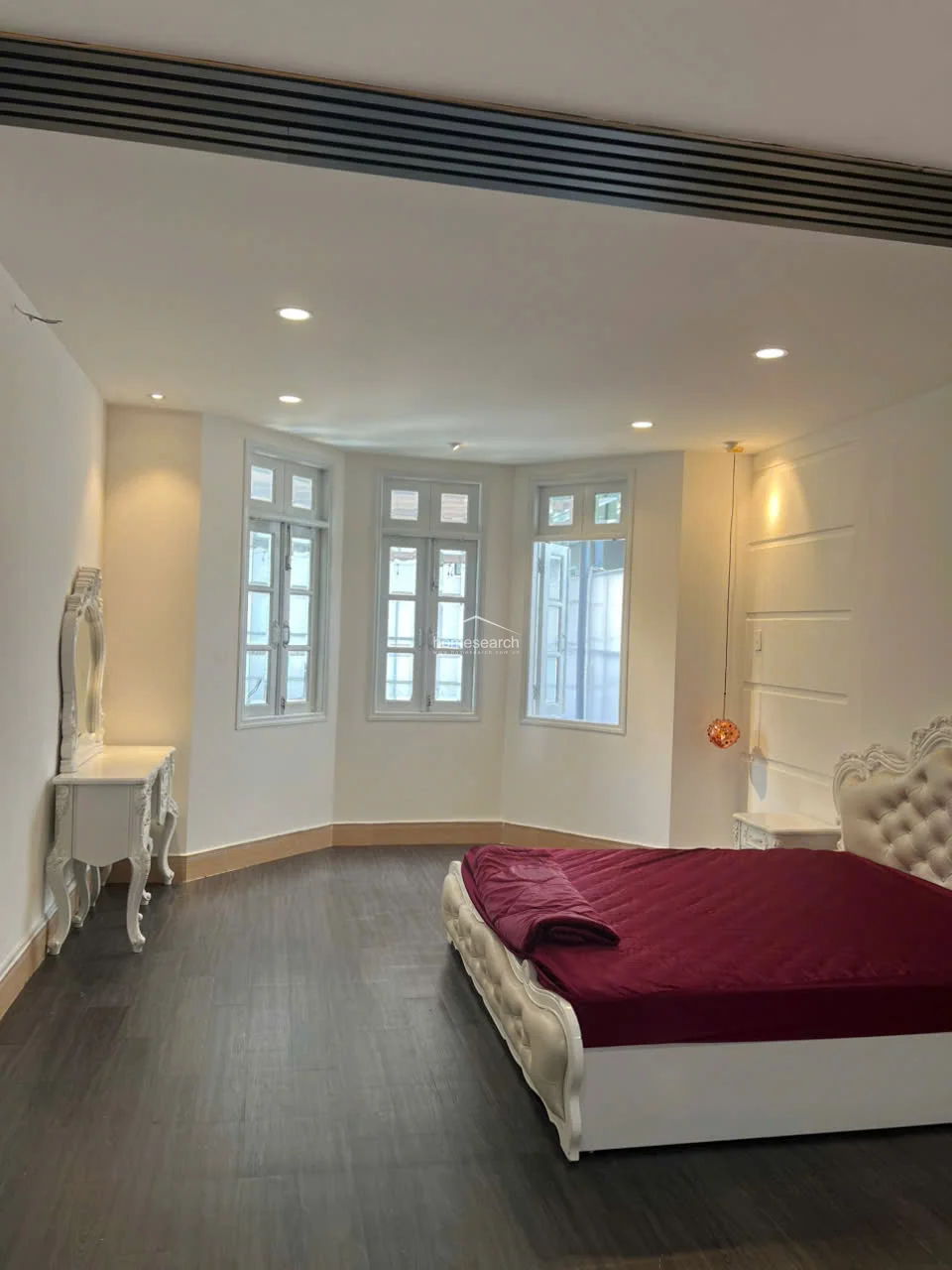 This spacious bedroom is defined by its dark wood-look flooring and a prominent bay window area that invites ample natural light. The room is anchored by an ornate, tufted cream-colored bed frame with deep red bedding, complemented by a matching vintage-style vanity and minimalist recessed ceiling lighting.