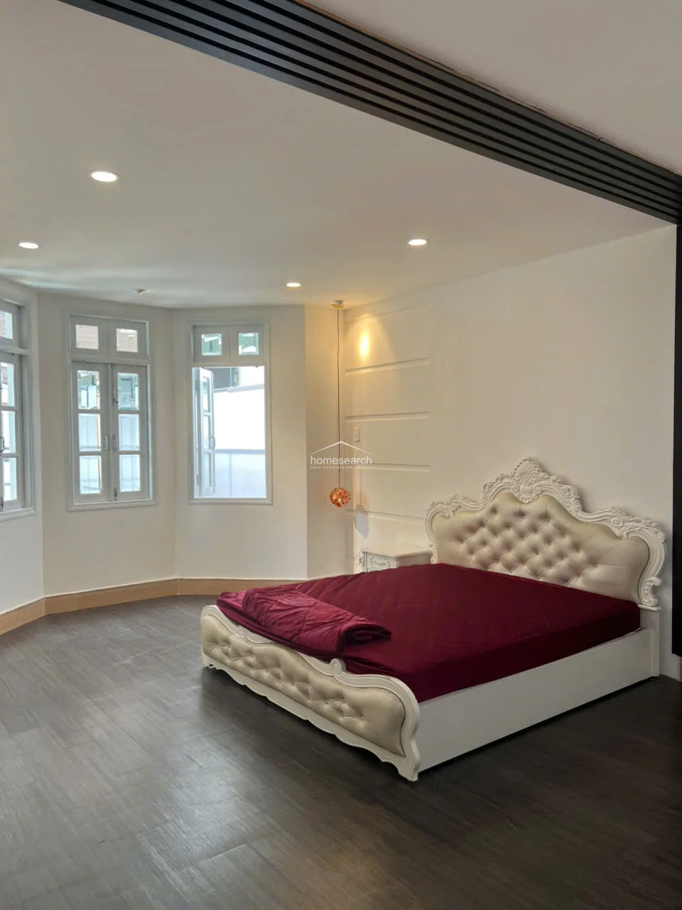 This bedroom maintains a consistent design language with the previous image, highlighting the elegant tufted headboard and the clean, white-walled aesthetic. The layout emphasizes the bay window as a focal point, creating a bright and airy environment that feels both luxurious and well-maintained.