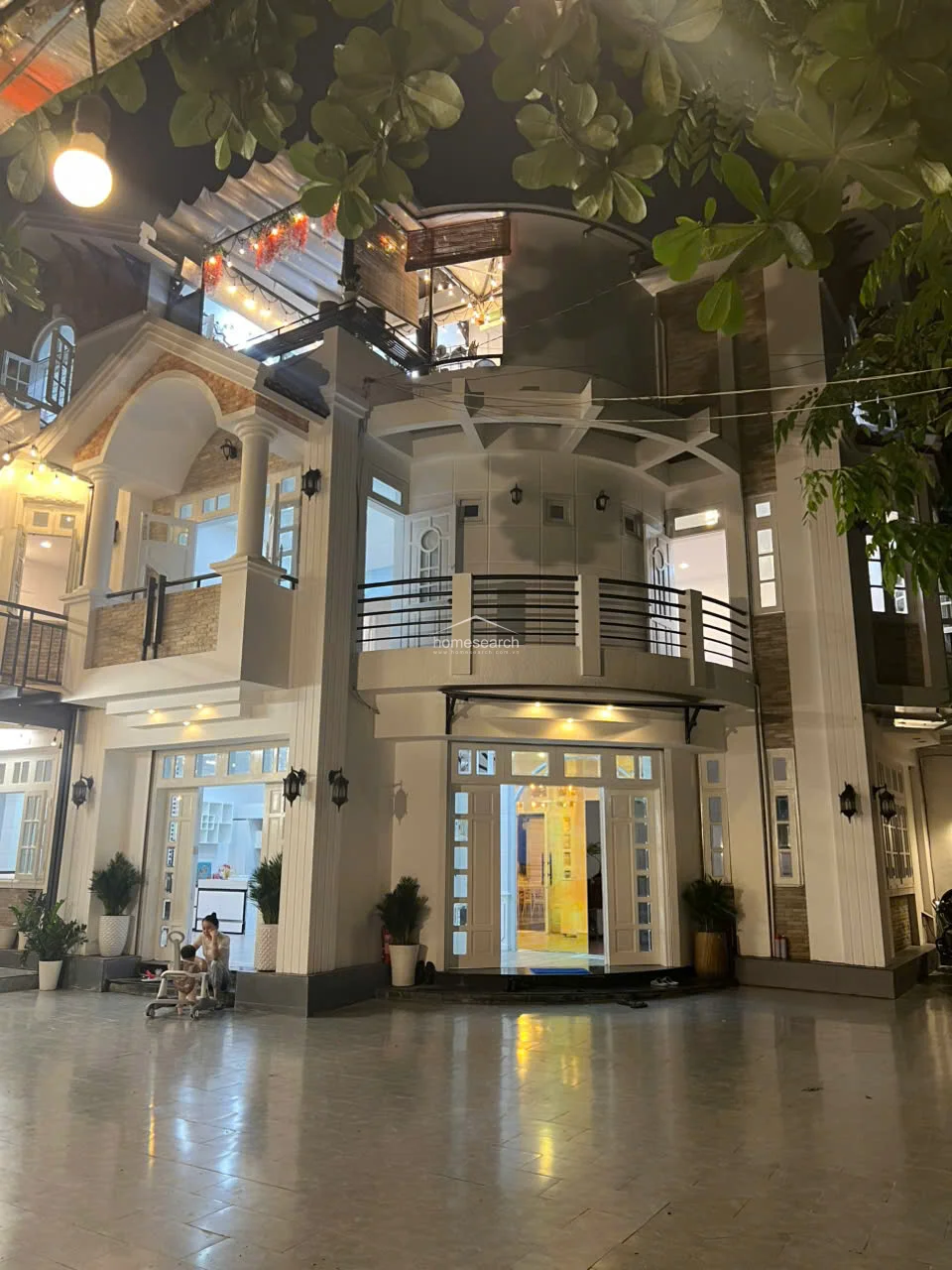 This striking exterior view showcases the property's sophisticated architectural details, including curved balconies, stone accents, and tiered levels. The home is well-lit with warm exterior lighting, highlighting the seamless transition between the indoor living areas and the large, open-air courtyard space.