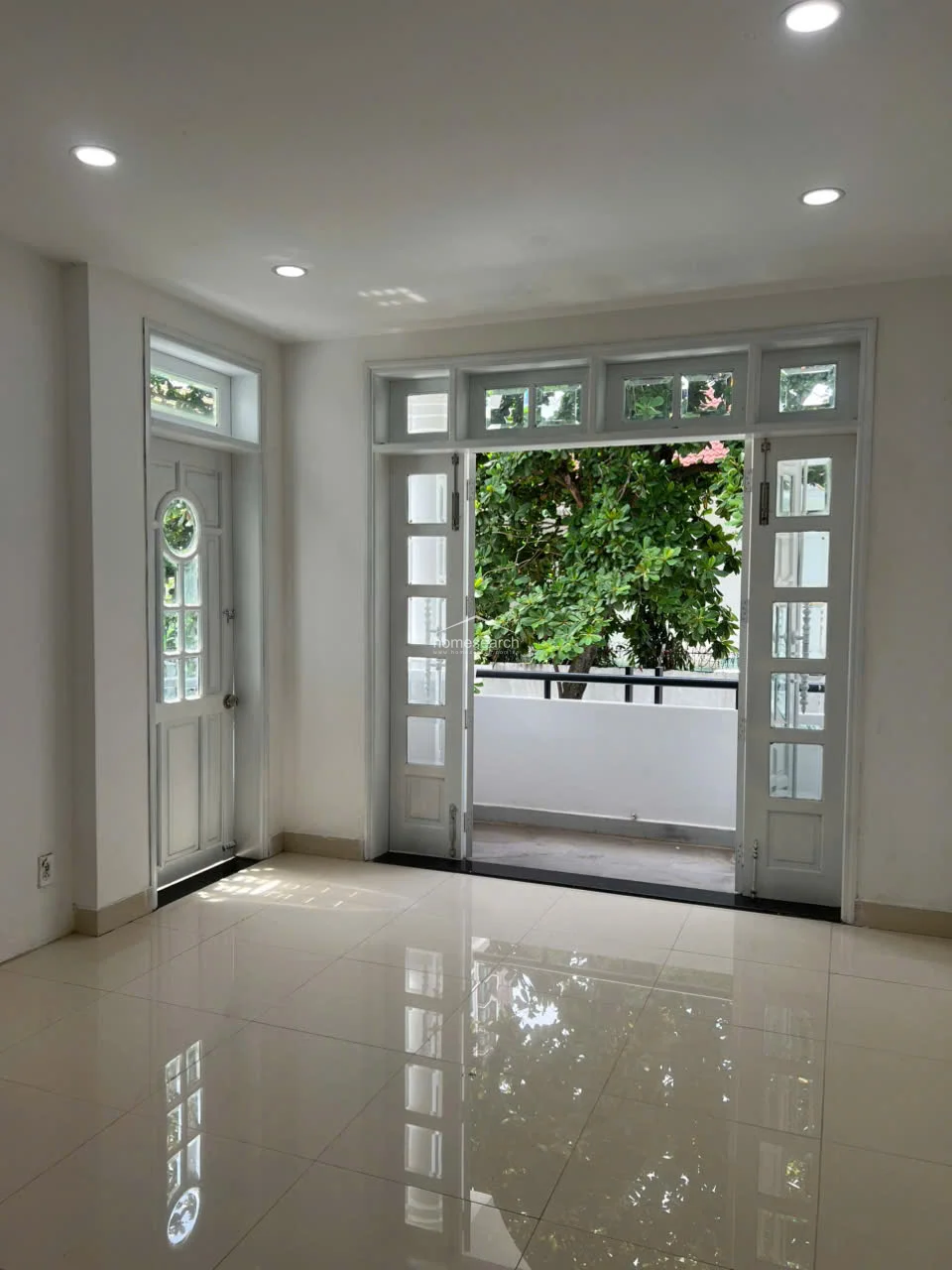 This bright, minimalist room features high-gloss white tile flooring and crisp white walls, creating a clean and airy atmosphere. It includes a functional balcony with a view of lush greenery, accessible through a large multi-pane glass door that allows for excellent natural light. The space is well-maintained with recessed ceiling lighting and a classic, architectural door design.