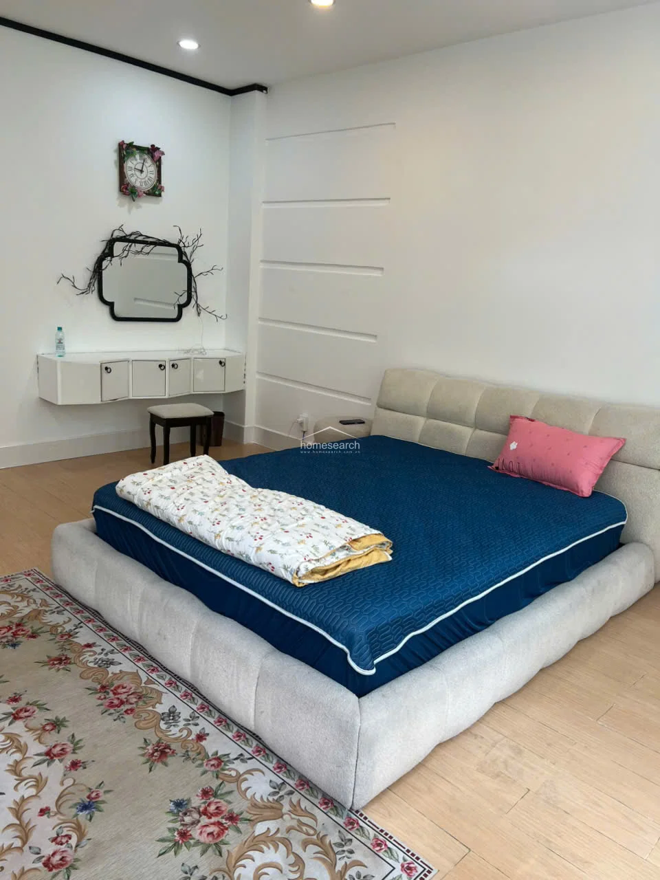 This bedroom offers a cozy and modern aesthetic with light-toned wood flooring and a neutral color palette. It features a contemporary upholstered bed frame, a floating white vanity with storage, and a decorative wall mirror. The room is well-lit and organized, providing a comfortable and tidy living environment.