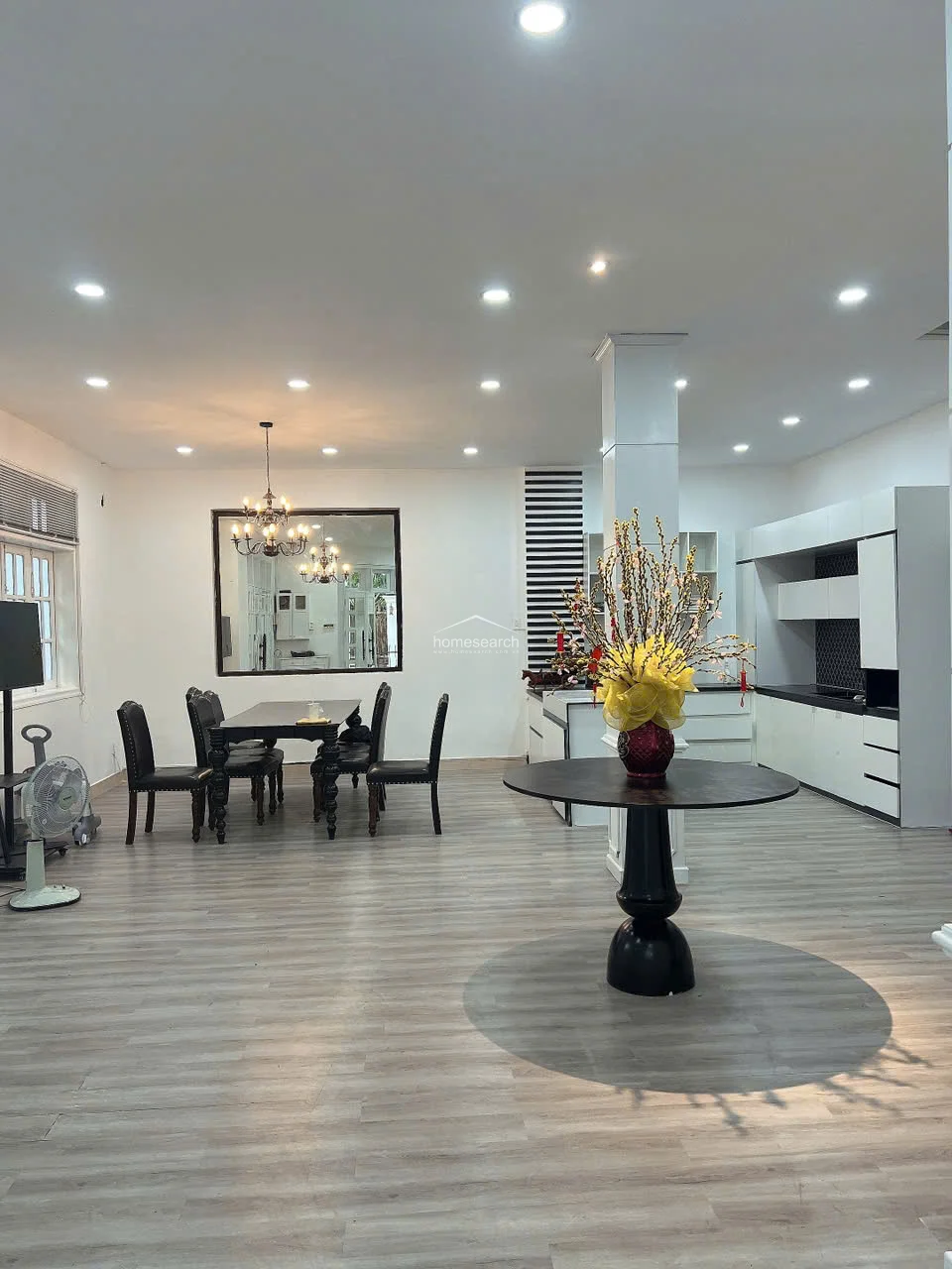 This expansive open-concept area seamlessly integrates the dining and kitchen spaces, featuring light grey wood-look flooring and a modern, monochromatic color scheme. The kitchen is equipped with sleek white cabinetry and a contrasting black backsplash, while the dining area is anchored by a sophisticated chandelier and a large wall mirror. A central column adds an architectural element to the room, which is illuminated by numerous recessed ceiling lights.