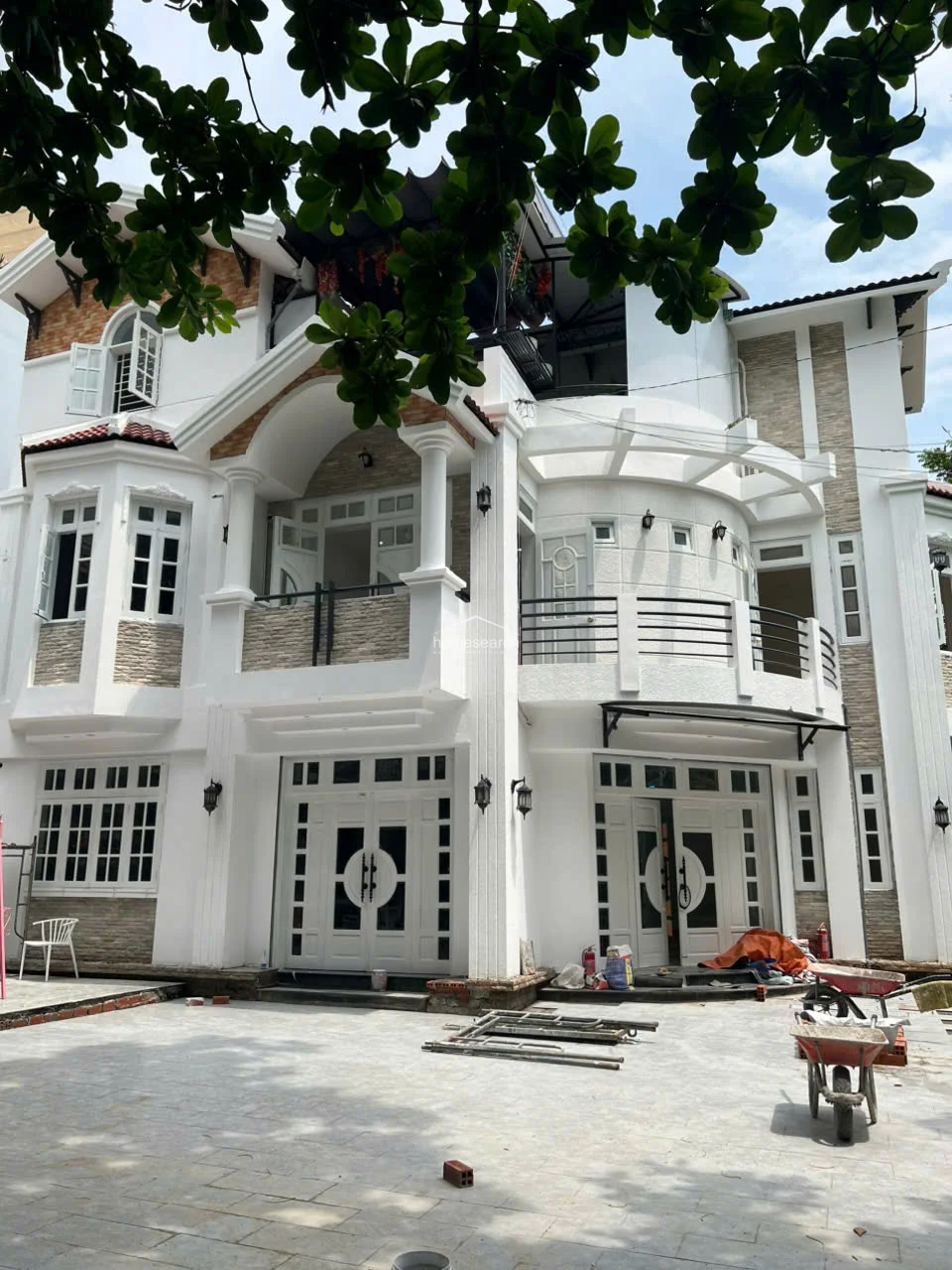This impressive multi-story villa showcases a grand architectural design with white stucco walls, stone accents, and classic columns. The exterior features multiple balconies, arched detailing, and large, ornate white doors, suggesting a high level of craftsmanship. While there is some ongoing construction equipment present, the overall structure appears well-maintained and provides substantial outdoor space.