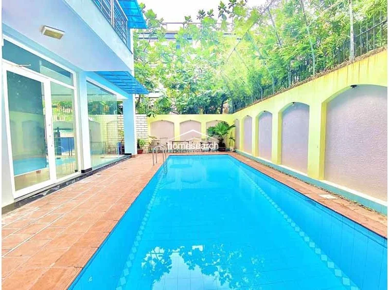 This image captures a long, rectangular swimming pool set against a privacy wall with decorative arched niches and lush greenery. The pool deck is finished with terracotta-toned tiles, creating a bright and inviting outdoor leisure space that connects directly to the indoor living areas through floor-to-ceiling glass doors.