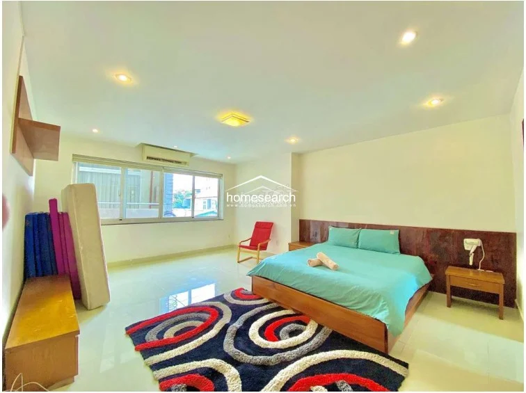 This bedroom maintains a clean, minimalist aesthetic with polished tile flooring and a prominent dark wood headboard wall feature. The space is equipped with a wall-mounted air conditioning unit, a vibrant patterned area rug, and a simple wooden nightstand, all illuminated by a combination of recessed and flush-mount ceiling lights.