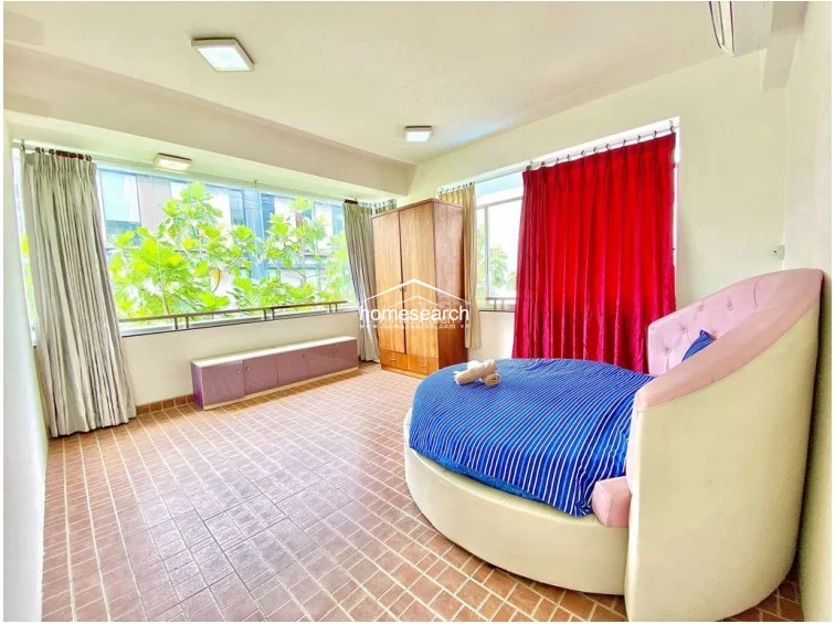 This bedroom features a distinctive circular bed with a pink upholstered headboard, set against a backdrop of vibrant red floor-to-ceiling curtains. The room includes a standalone wooden wardrobe and provides direct access to a balcony through large glass doors, allowing for plenty of natural light and ventilation.