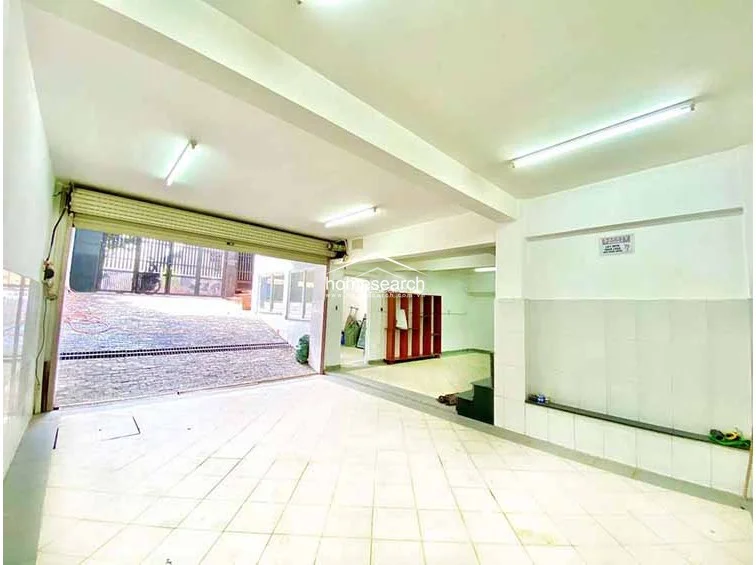 This interior view of the garage or utility space shows a clean, minimalist layout with large-format light-colored floor tiles and white walls. The area is well-lit by overhead fluorescent lighting and features a roll-up metal door, suggesting a secure and functional space for vehicle storage or additional multipurpose use.