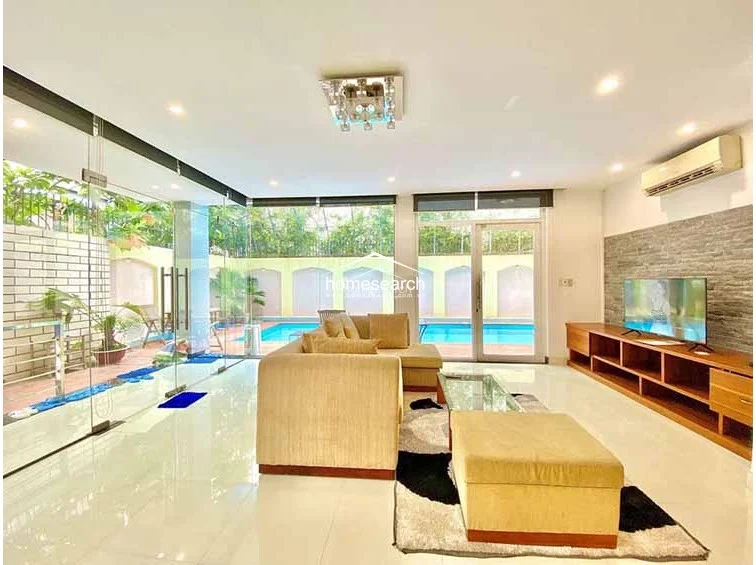 This living room features a bright, open-plan layout with polished light-colored flooring and a feature wall clad in textured stone. The space is furnished with a neutral-toned sofa and a wooden media console, while floor-to-ceiling glass walls offer an unobstructed view of the pool, creating a sophisticated and airy atmosphere.